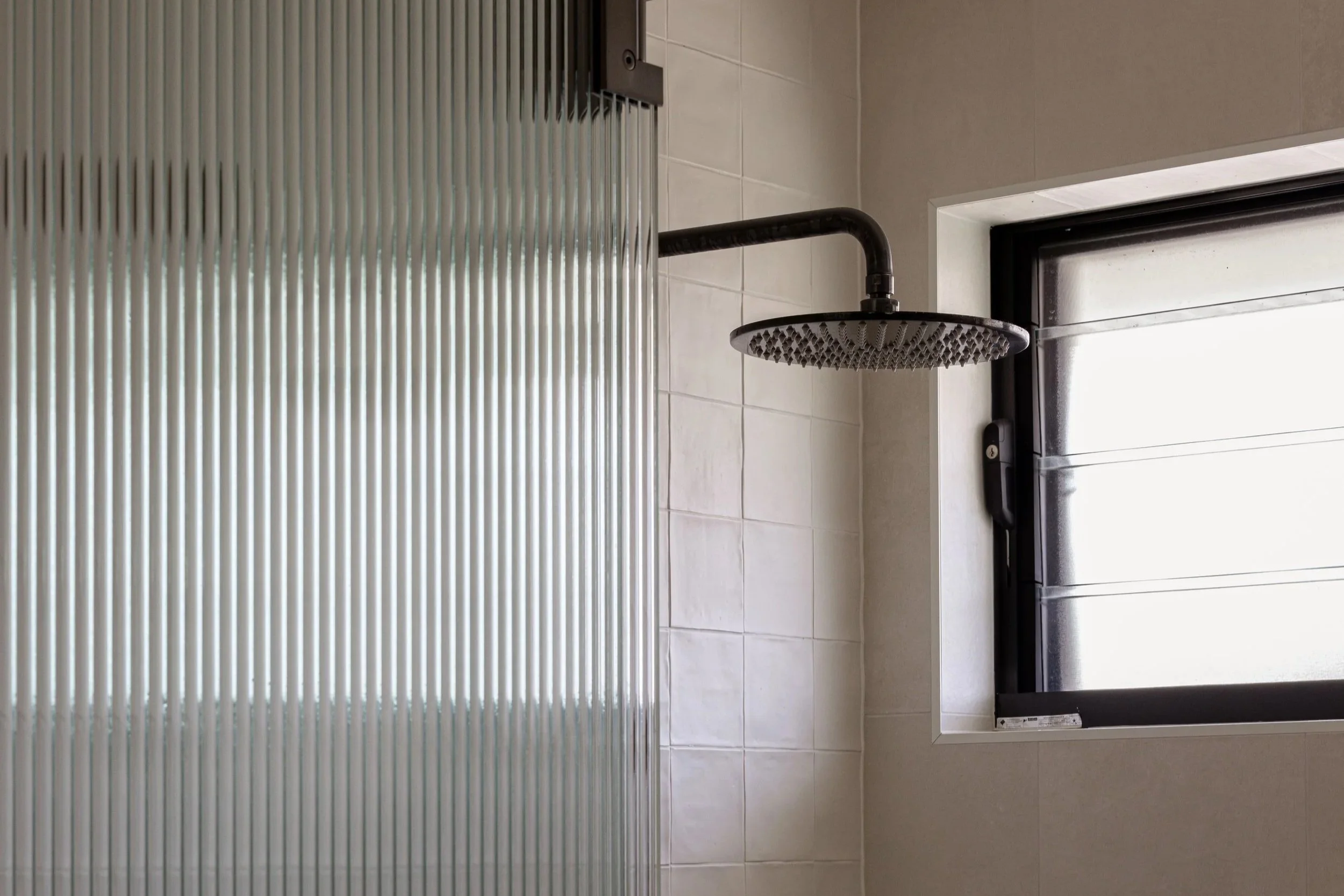 Shower area with a black showerhead and a partially frosted window, tiled walls, and a sliding glass door with vertical ridges.