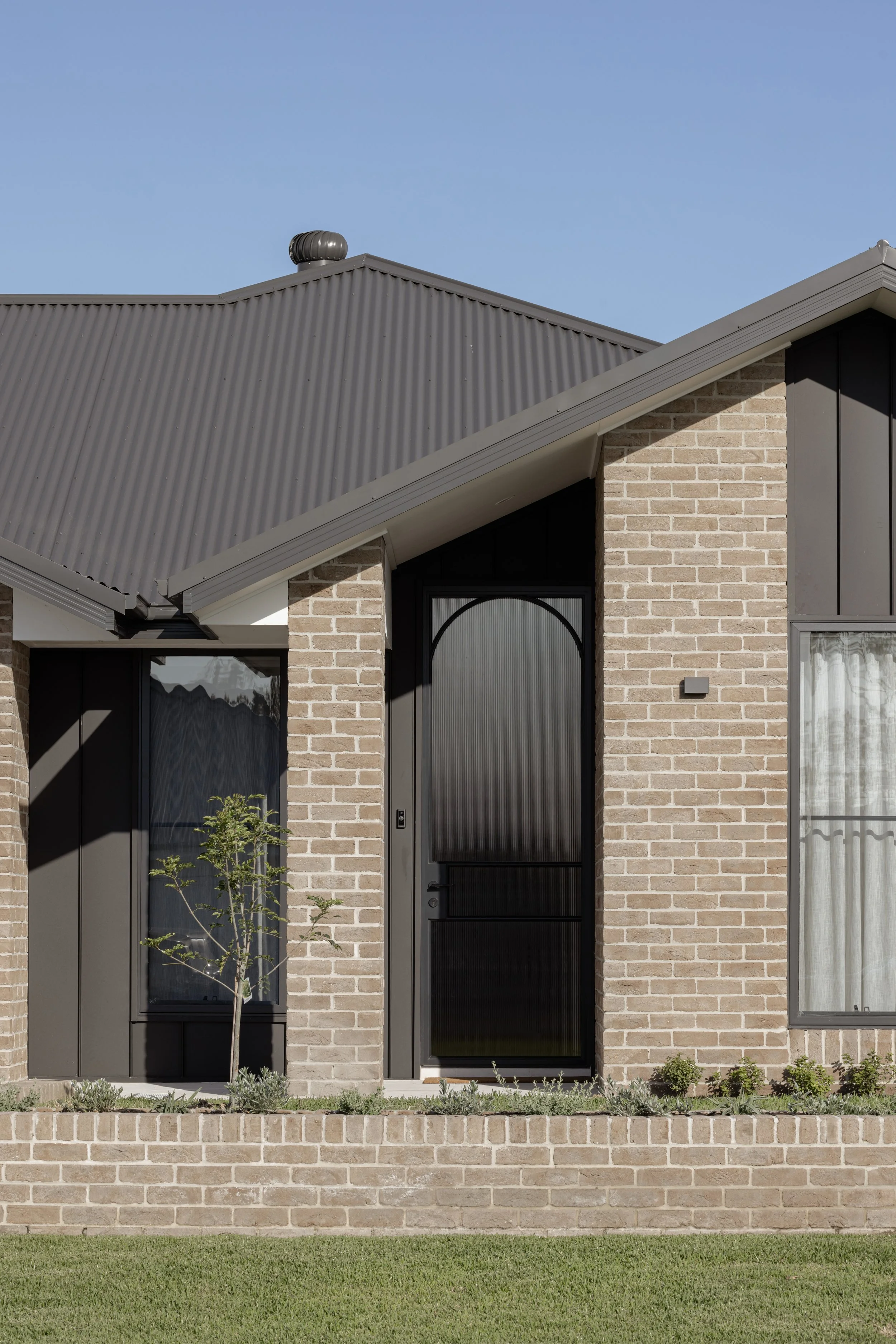Front view of a modern house with brick walls, black entry door, large windows with curtains, small trees and plants in the garden, and a dark gray metal roof under a clear blue sky.