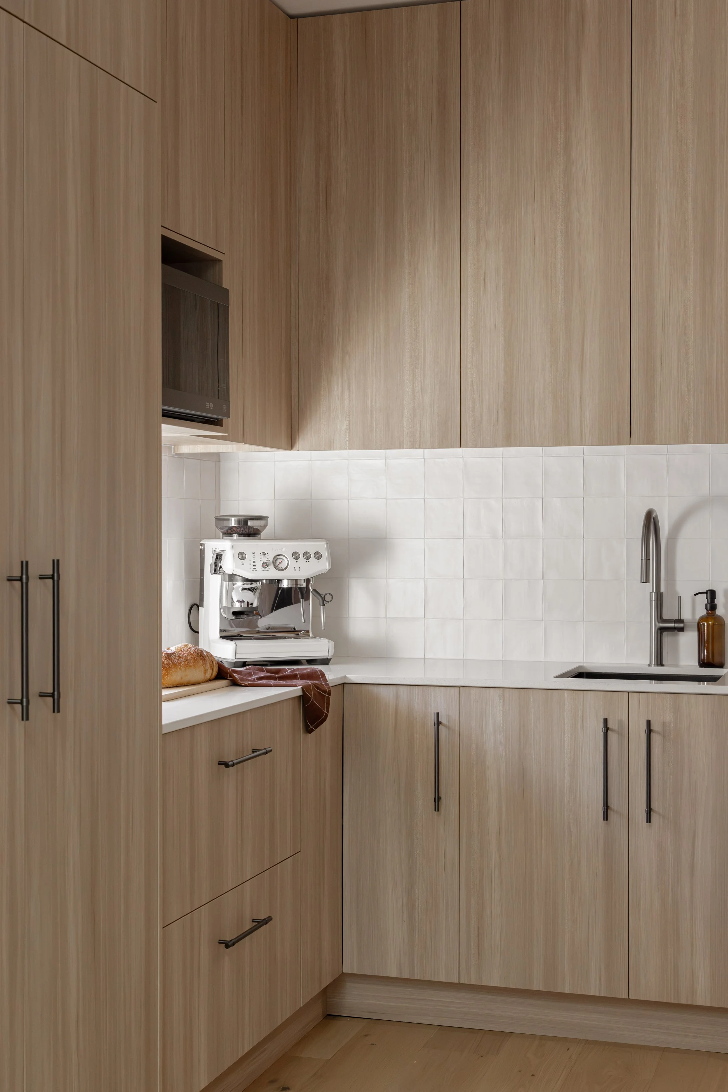 A minimalist kitchen with beige wood cabinets, a white tiled backsplash, a coffee machine on the counter, a loaf of bread, and a brown cloth. There is a stainless steel sink with a soap dispenser and a modern faucet.