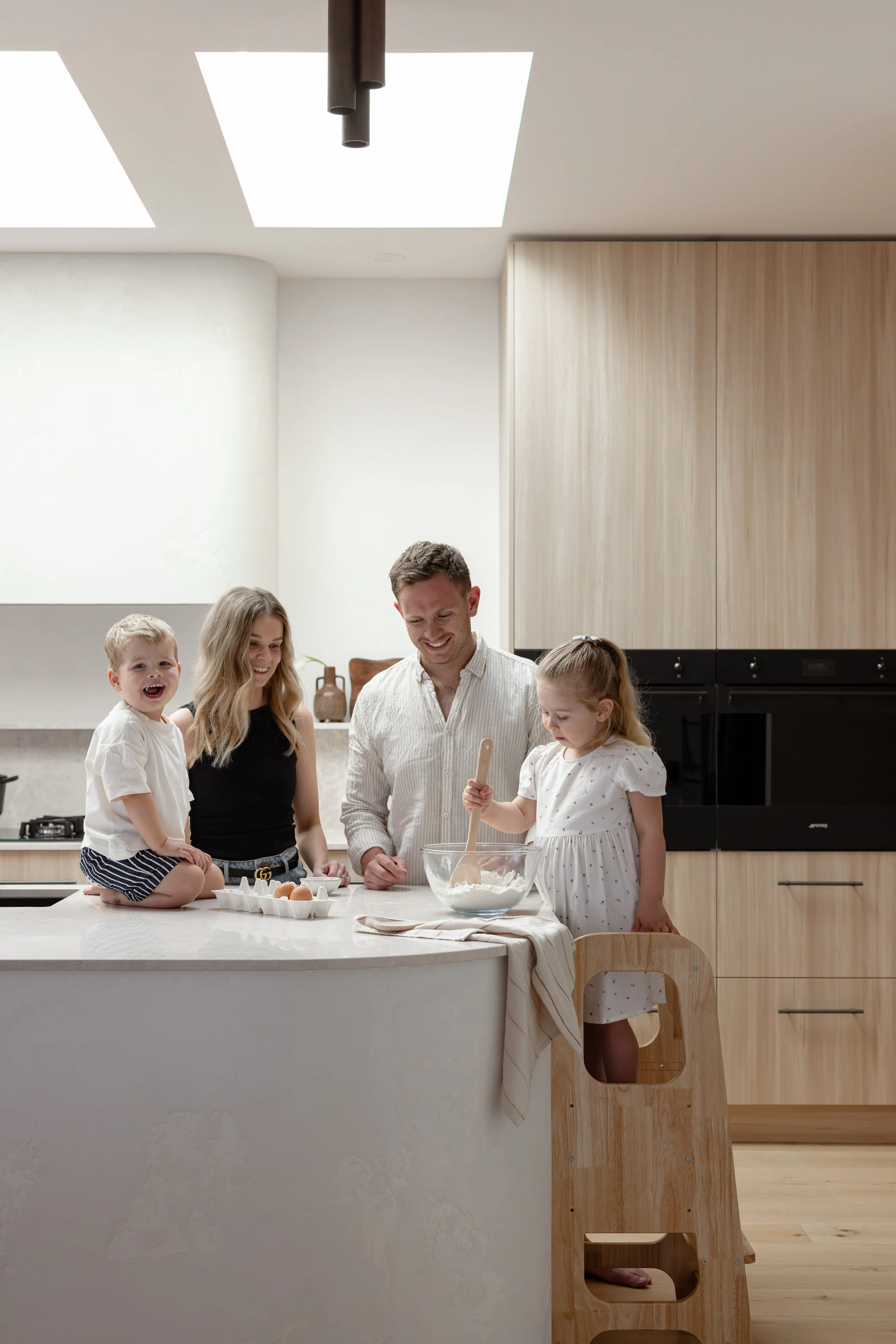 A family of four, including two young children, is gathered around a kitchen island. They are baking together, with one child mixing ingredients in a bowl, while the others watch and smile. The kitchen is modern with light wood cabinets and a large window providing natural light.