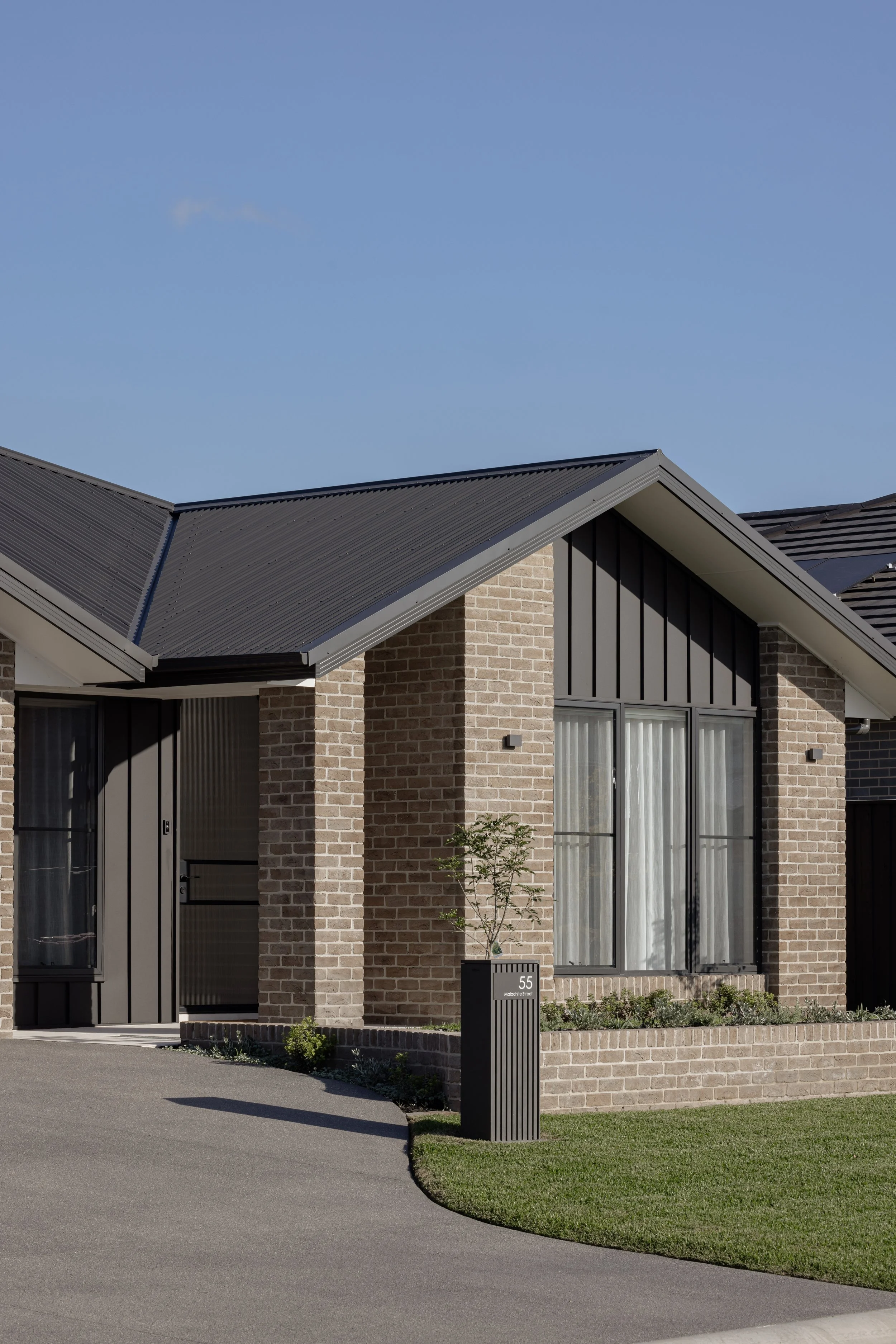 Modern house with brick exterior, black window frames, and a gray metal roof. A pathway curves alongside a grassy lawn with a small tree in front and a black mailbox with the house number 55.