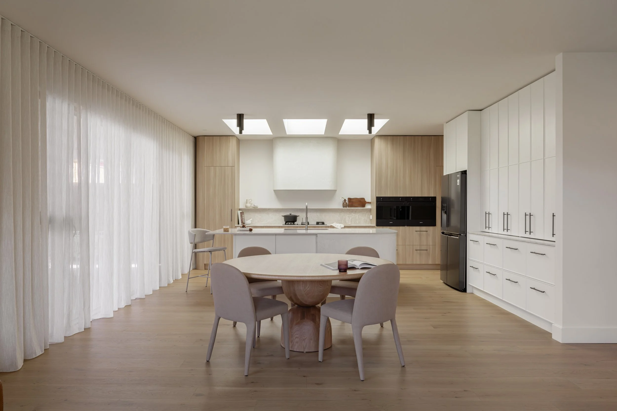 Modern kitchen with wooden cabinets, white countertops, round dining table with four chairs, and large sheer curtains on windows.