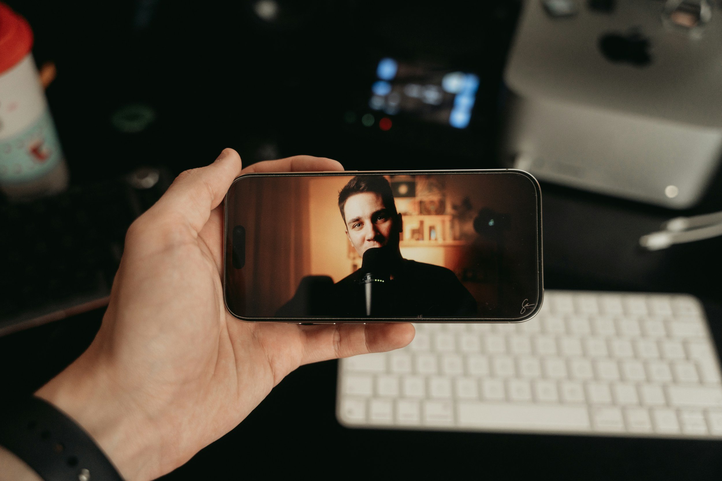 Person holding a smartphone capturing a selfie of a young man with a microphone in a dimly lit room.