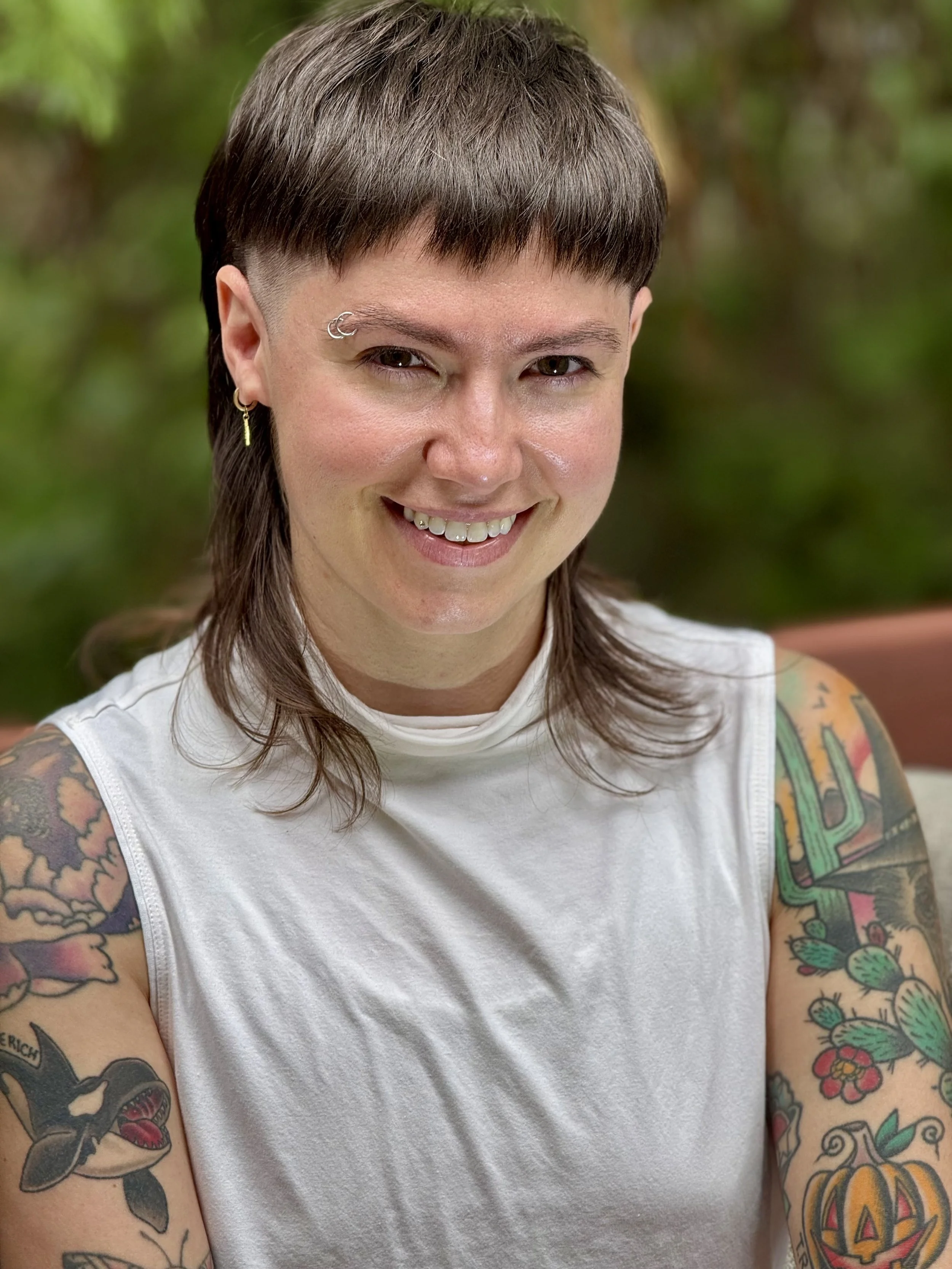 Genderqueer person with a mullet and brown hair, eye brow rings, earrings, and a tooth gem in a white tanktop sits in front of a blurred wooded background. This person has many visible tattoos depicting plants and animals
