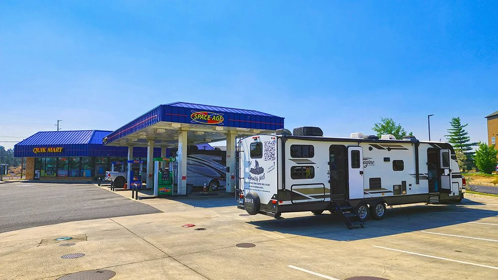 An RV travel trailer sits in the foreground with both doors open.  In the background is a Space Age gas station.  On the front of the building with blue awnings, it says Quick Mart.  There are various trucks and RVs at the gas pumps.
