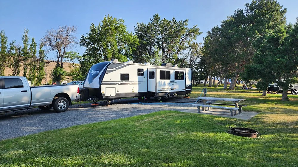 A Grand Design 3000QB is parked in a camping spot at Wanapum Recreation Area.  There is green grass, a firepit and picnic bench in the foreground and trees in the background.
