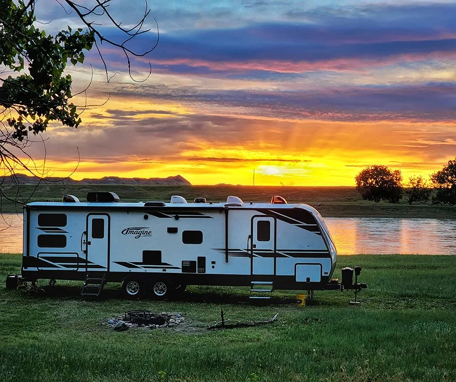 A breathtaking picture of a Grand Design 3000QB sitting on green grass with a river in the background and the rising sun with yellow and pink sky.