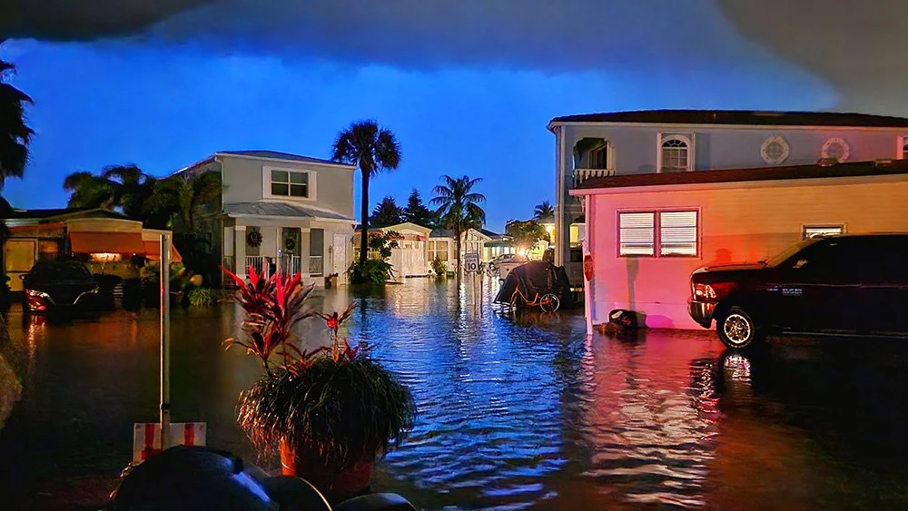 A picture taken on a corner of a very flooded Florida community at night.  Various shades of dark blues are in the sky and reds reflecting off a nearby home.