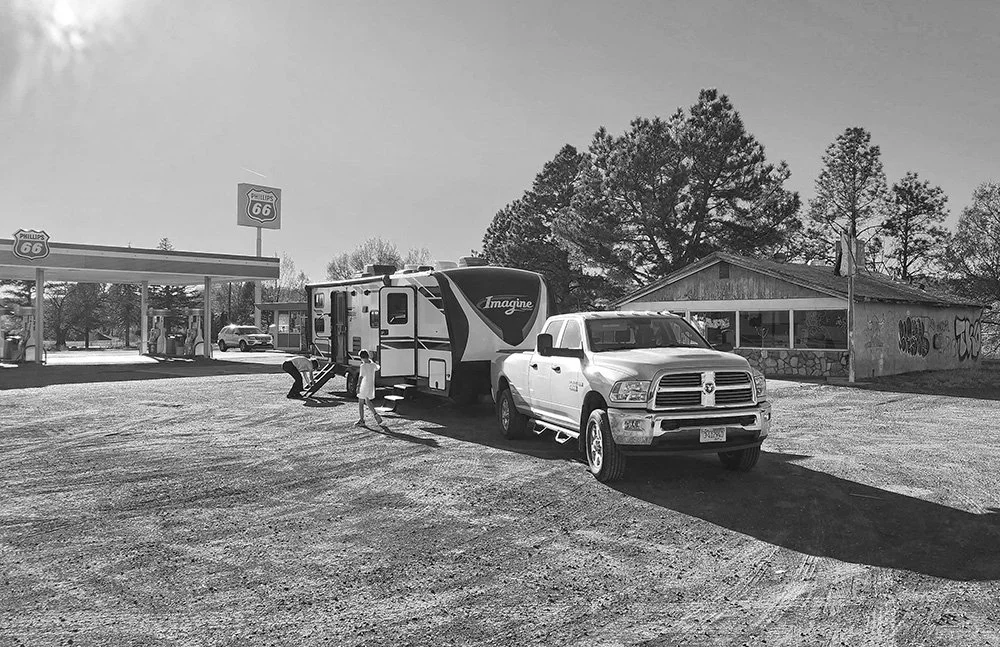 A black-and-white photo of a truck and RV with a fuel station in the background and an old, abandoned building to the right.