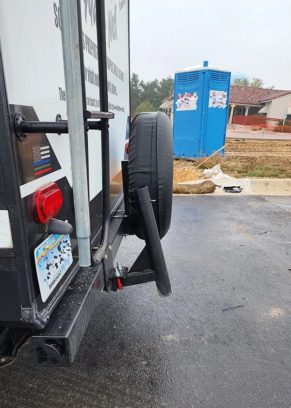 A picture of the rear of an RV from the side view shows the flagpole hitch accessory bent upwards.  A blue outhouse is in the background.