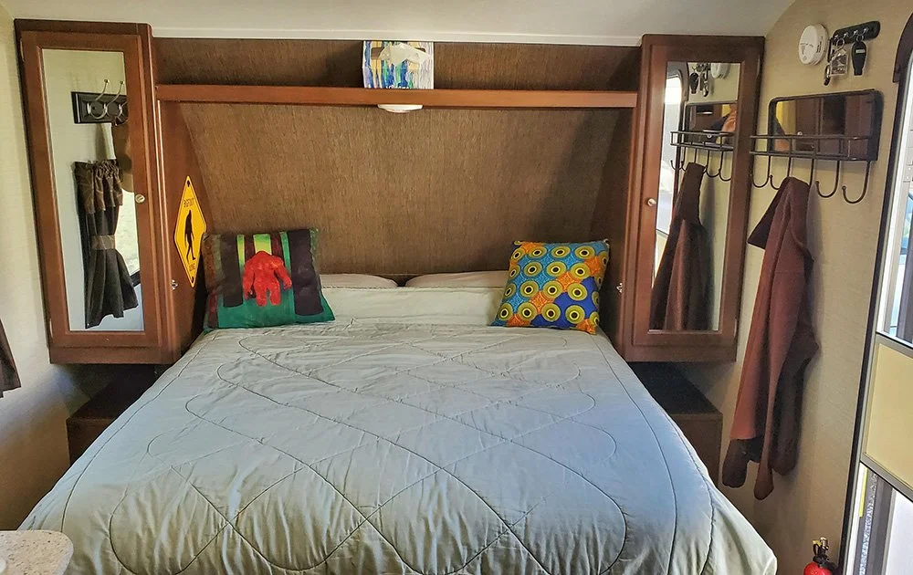 The interior of an RV focusing on a murphy bed.  Tow pillows lay on the bed: one of a Sasquatch and the other of a colorful design of yellow bursts on blue, orange, and dark blue stripes.