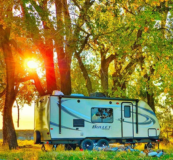 A Keystone Bullet travel trailer sites in a picturesque grassy fields with large cottonwood trees in the background.  The golden setting sun peaks through the trees.