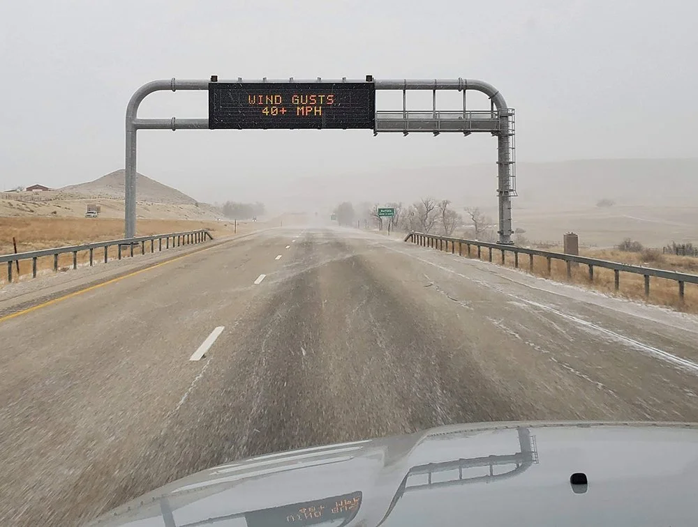 A POV of a vehicle driving down a snow-dusted interstate with an overhead sign in the foreground saying WIND GUSTS 40+ MPH.