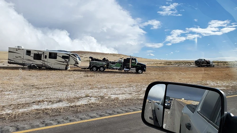 A crashed 5th wheel is hooked to a large tow truck in on the Interstate median.  The crashed truck is in the background on top of another flatbed tow truck.