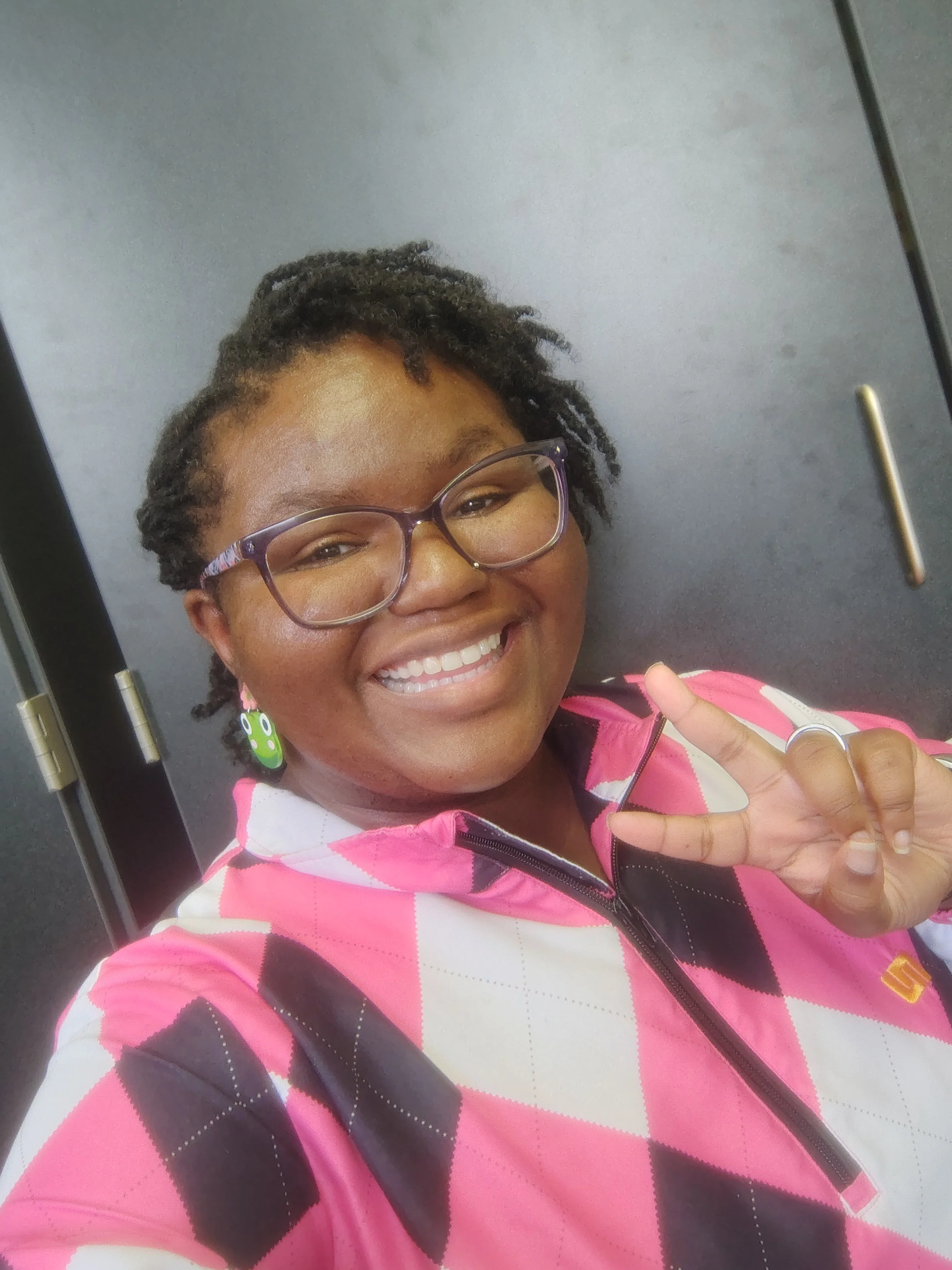 Smiling woman with glasses and earrings, making a peace sign with her hand, wearing a pink, black, and white patterned jacket, photographed indoors against a plain gray background.