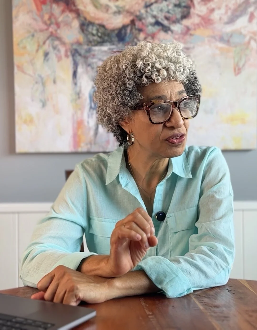 An older woman with curly gray hair and glasses sitting at a wooden table, wearing a light blue shirt, with a colorful abstract painting in the background.