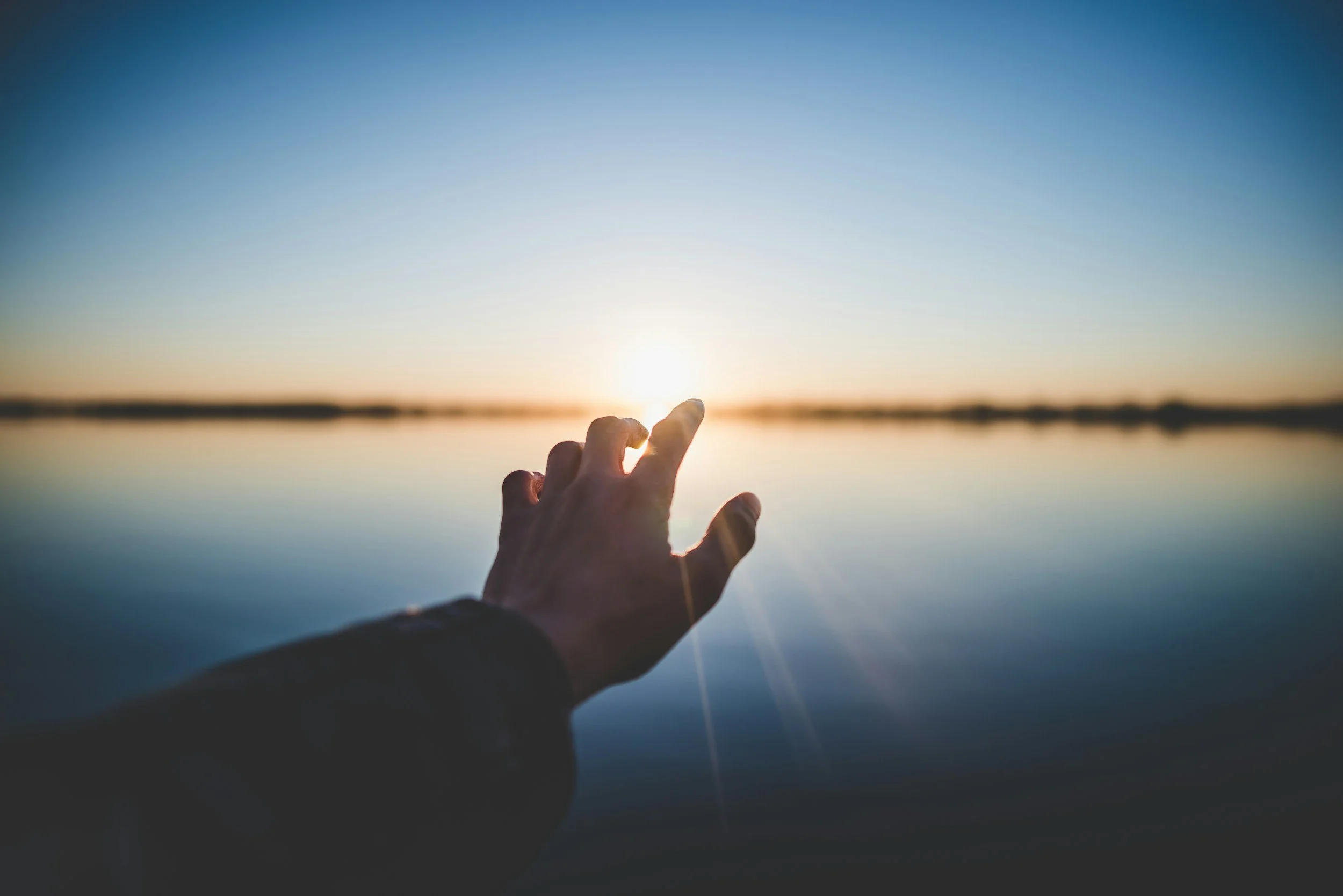 Hand reaching towards the setting sun over a calm body of water during sunset.