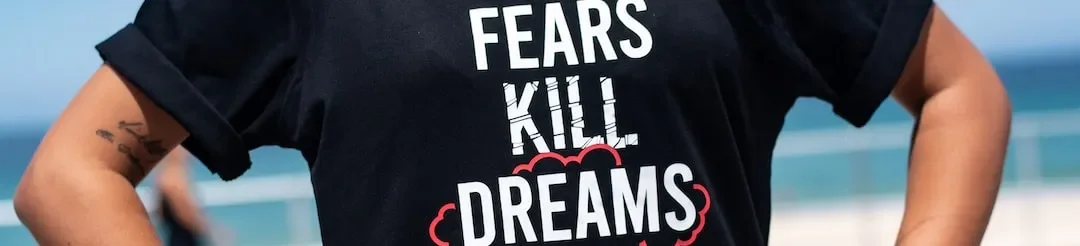 Person wearing a black t-shirt that says "FEARS KILL DREAMS" on a beach or waterfront area.