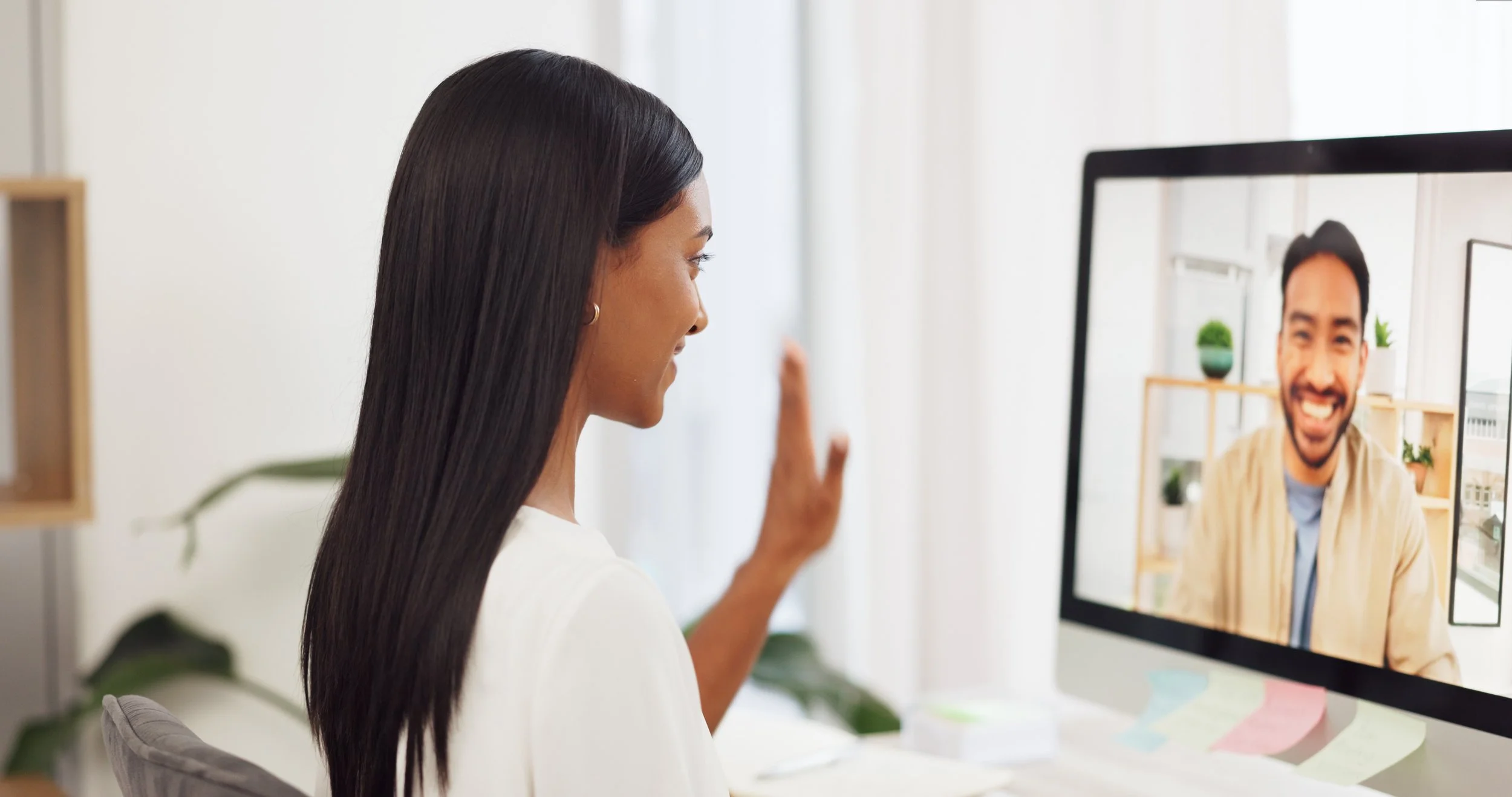 Woman video doing telehealth therapy on computer, waving hello to smiling man on screen.