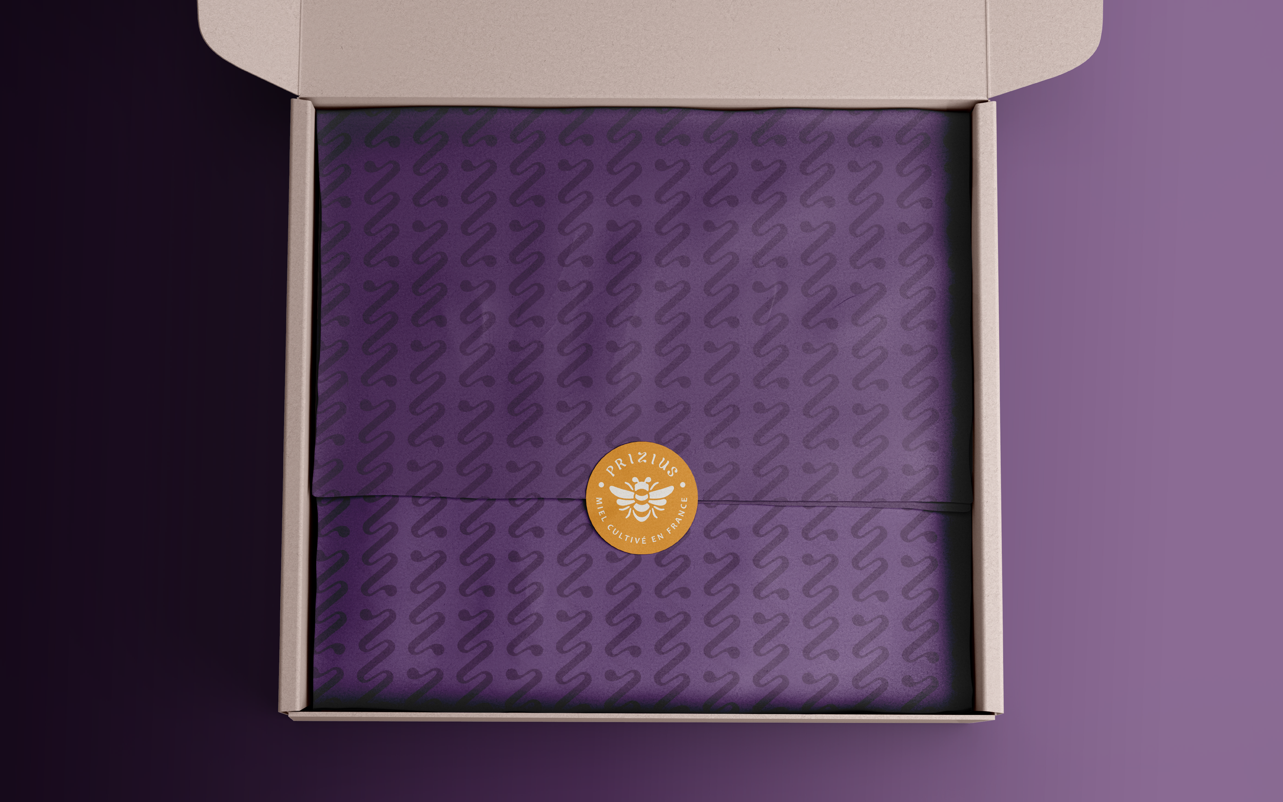 Une boîte en carton beige ouverte contenant une boîte en violet avec un motif en forme de serpent et un badge orange avec un abeille, texte en français, indiquant que le miel est cultivé en France.