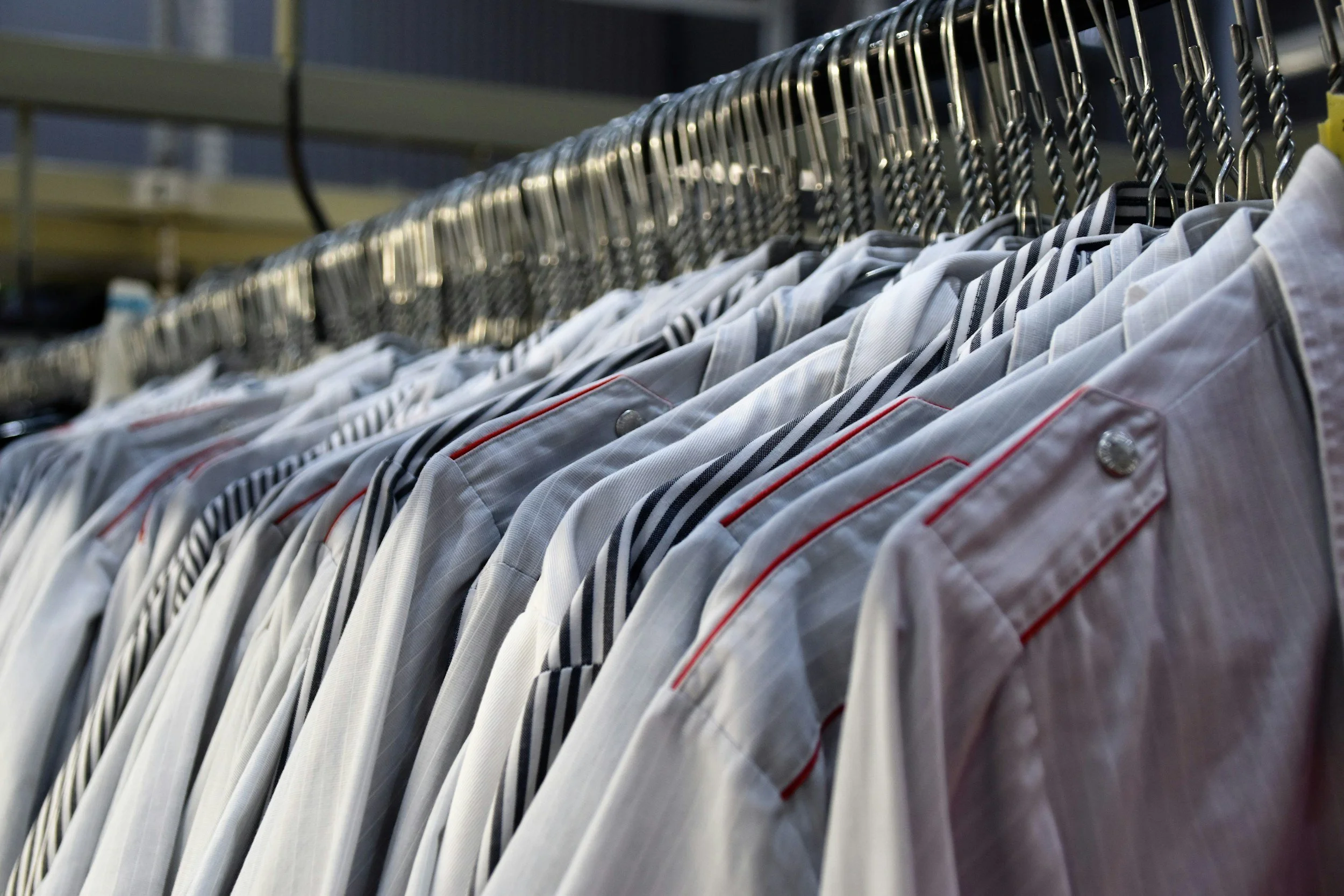 A row of white men's dress shirts hanging on a metal clothing rack, some with striped patterns and red piping.