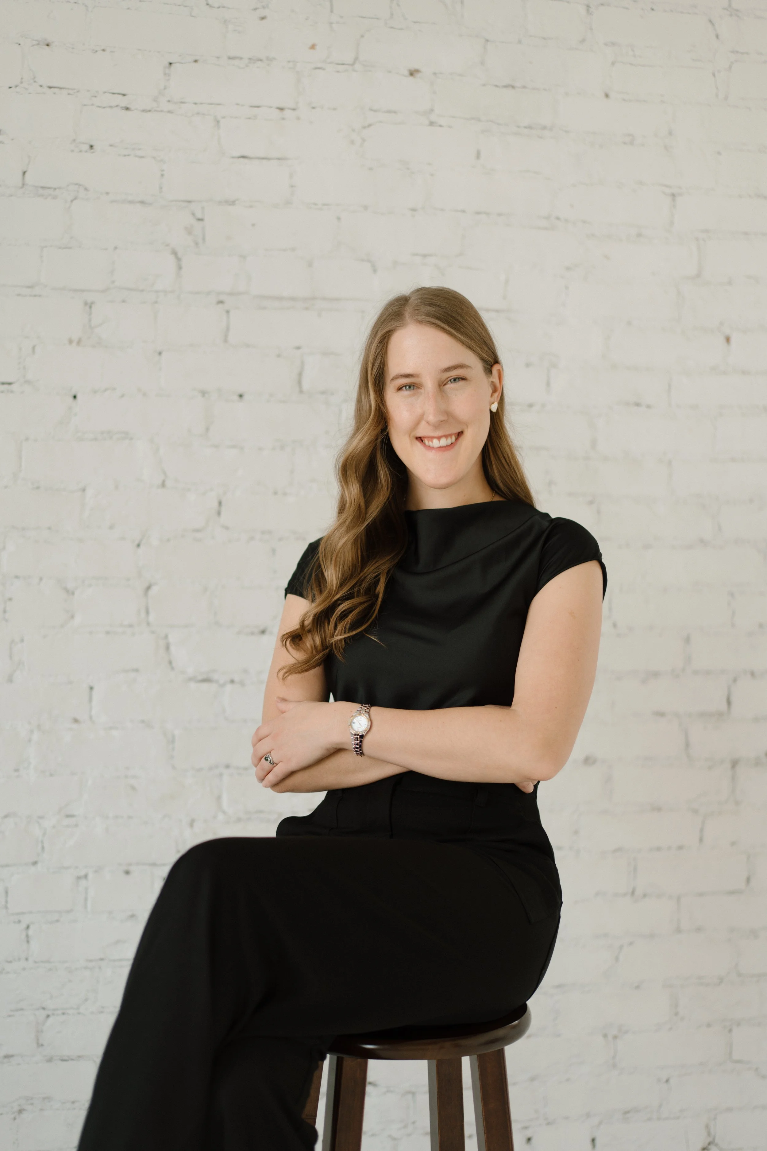 A confident young woman with long wavy brown hair, wearing a black outfit, sitting on a stool against a white brick wall, smiling with her arms crossed.