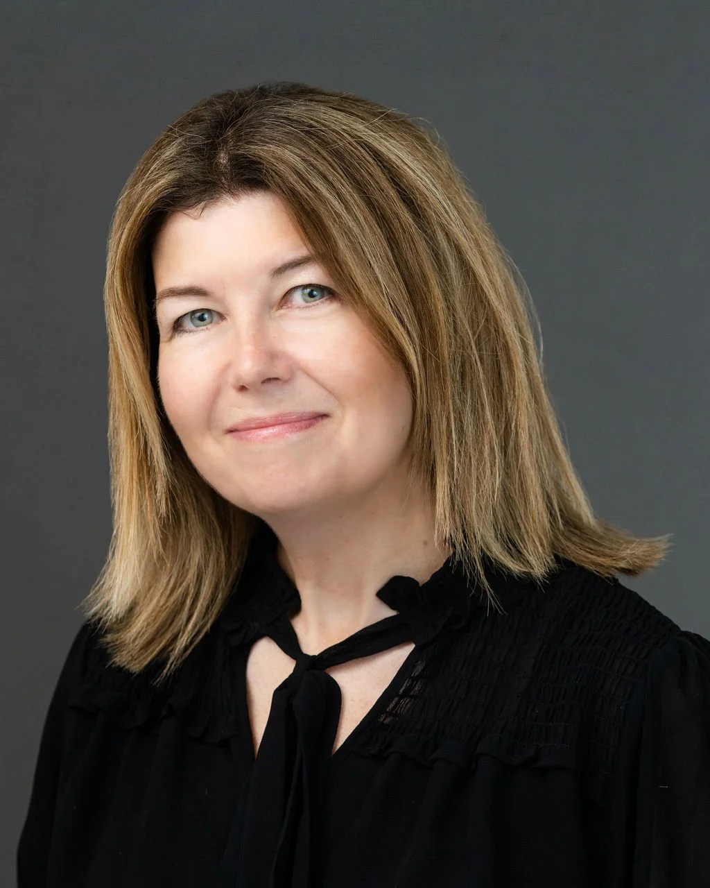 A woman with shoulder-length light brown hair and blue eyes, wearing a black blouse with ruffled and smocked details and a bow tie at the collar, against a dark gray background.