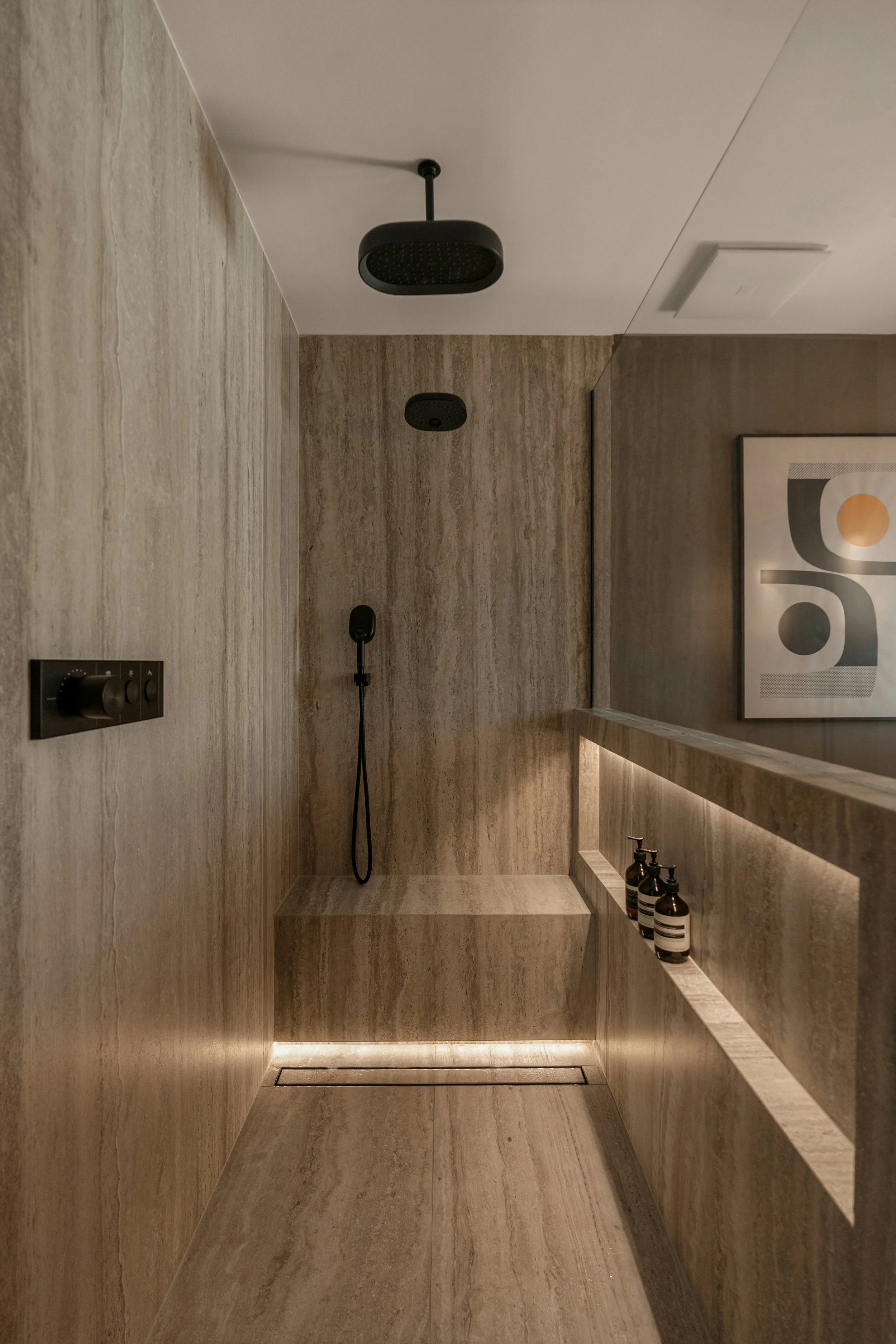 Minimalist bathroom shower area with beige marble walls and floor, overhead black showerhead, handheld shower, recessed lighting, and three bottles of soap or shampoo on the ledge.