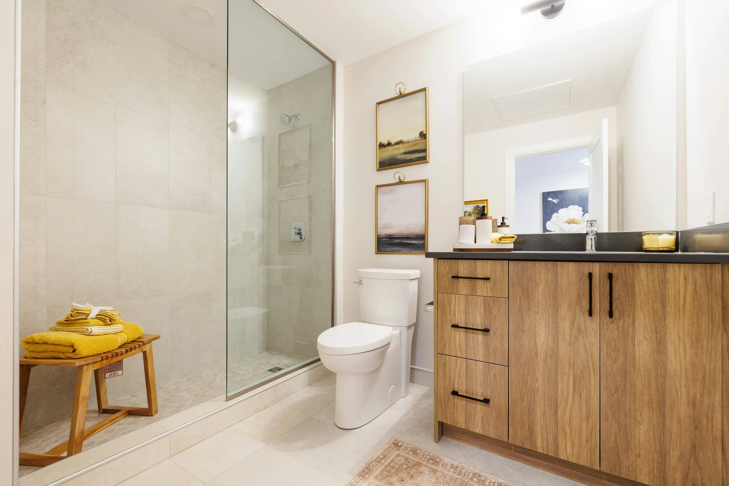 Modern bathroom with a glass-enclosed shower, a white toilet, and a wooden vanity with black handles. The vanity has a black countertop with toiletries and a large mirror. Decorative paintings hang on the wall above the toilet, and the bathroom has soft, natural lighting.