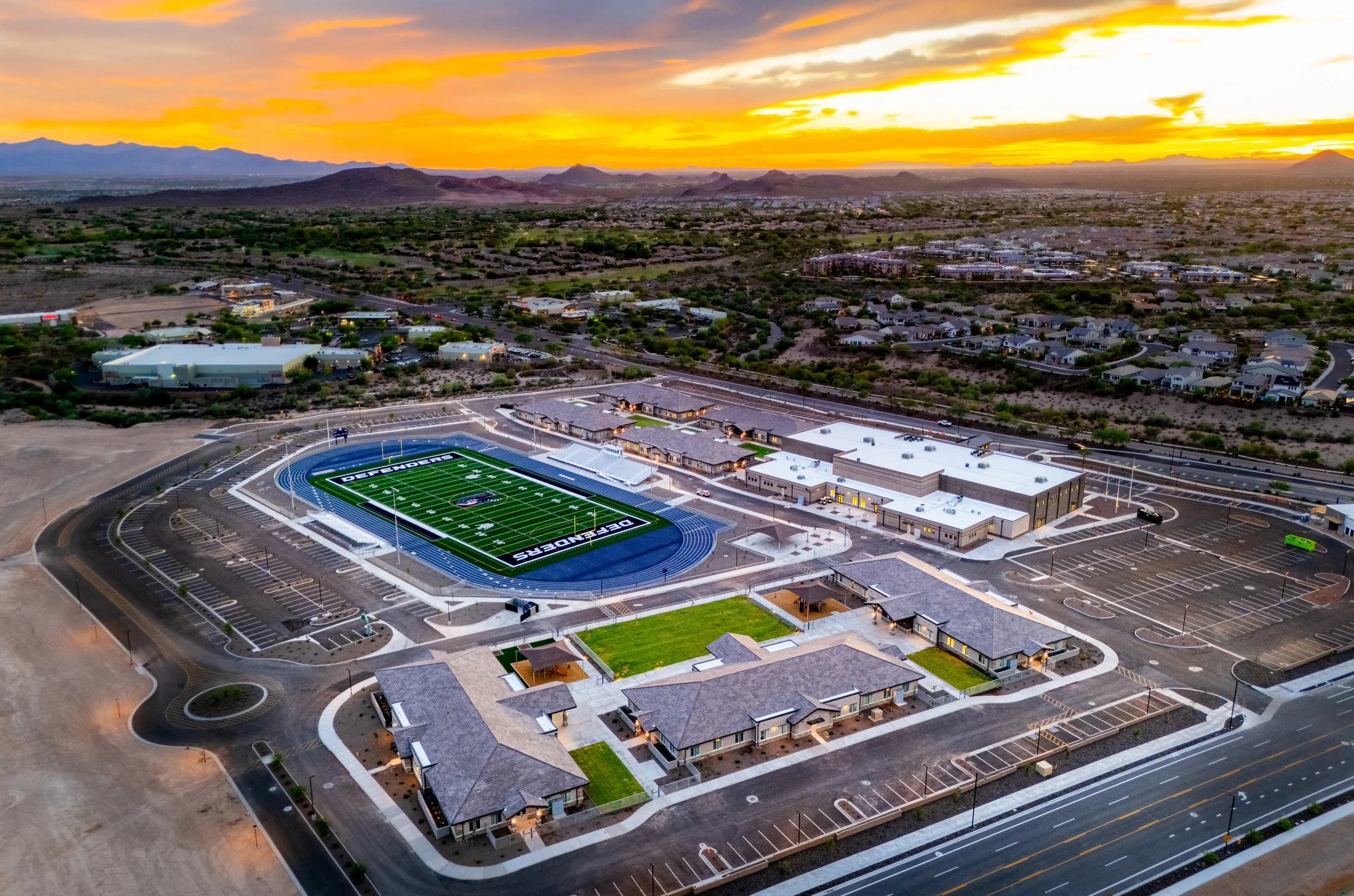 Aerial view of a new school campus featuring a football field with blue track, parking lots, and surrounding buildings at sunset, with mountains in the distance.