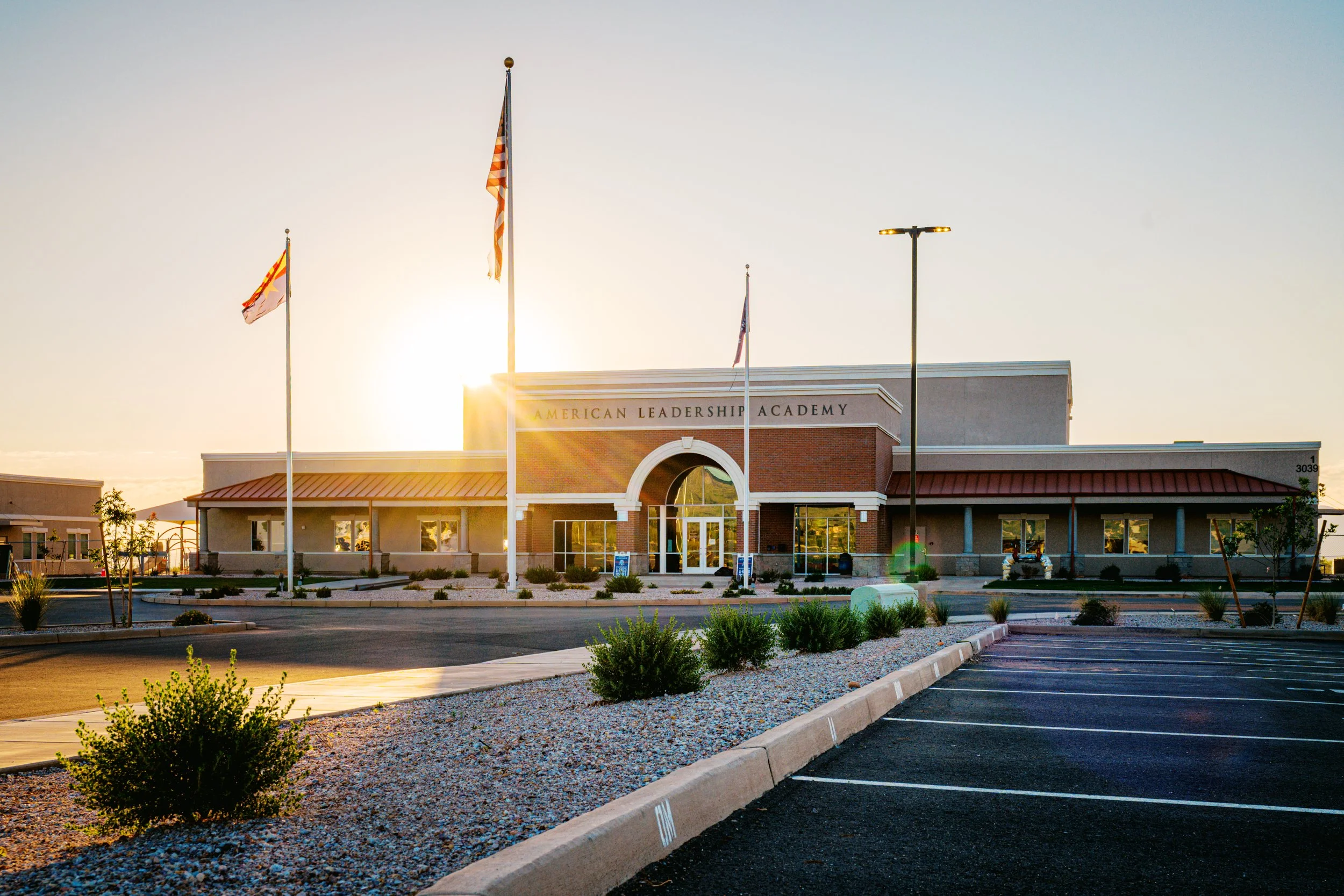 Front view of the American Leadership Academy building at sunset, with flags flying in the foreground and an empty parking lot in the front.