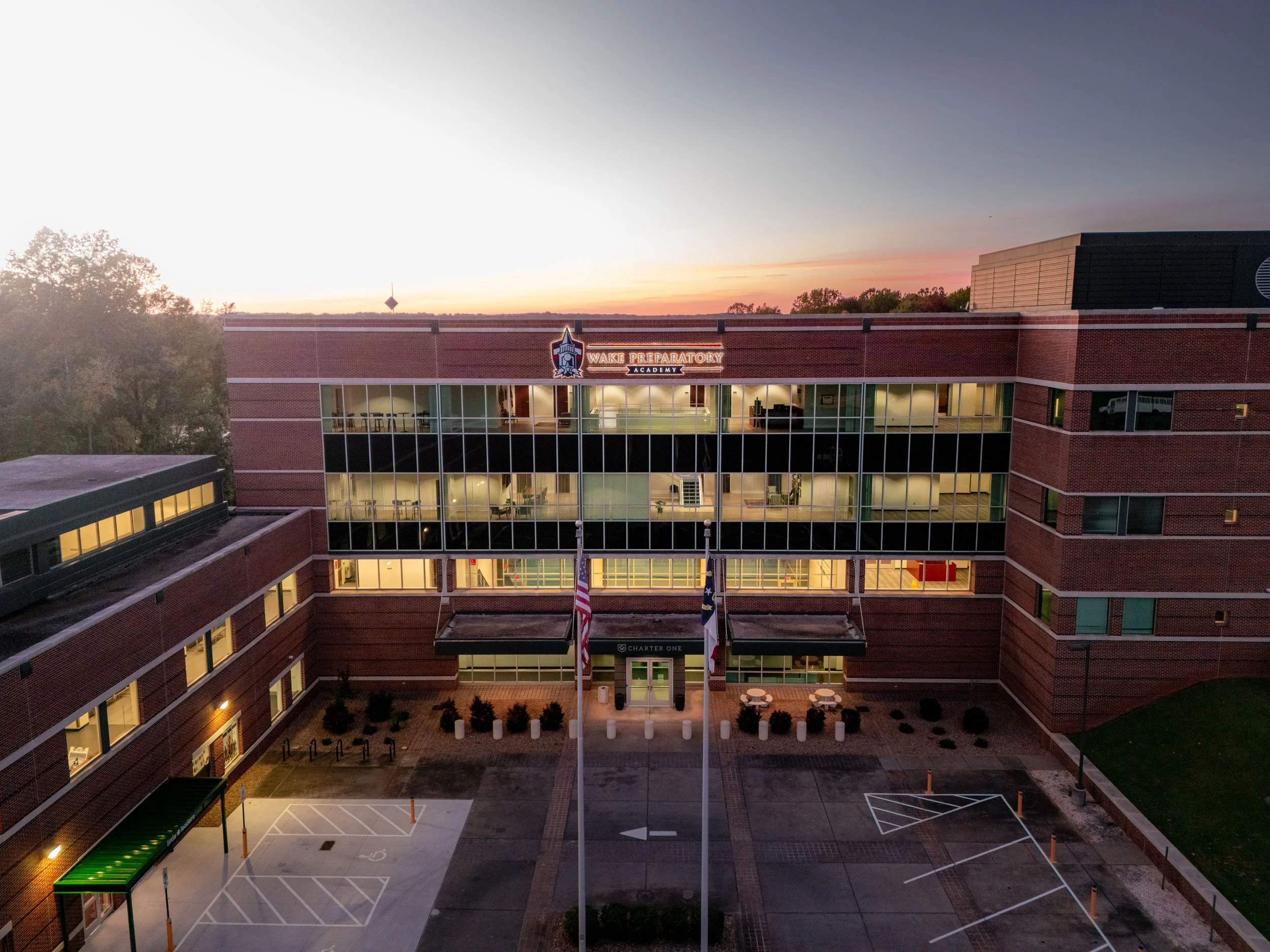 A large brick school building with glass windows, labeled 'Wake Preparatory Academy,' at sunset. The building has multiple floors, and there are flagpoles with American and Texas flags in front. The parking lot nearby is mostly empty, with a few marked parking spaces and a bike rack.