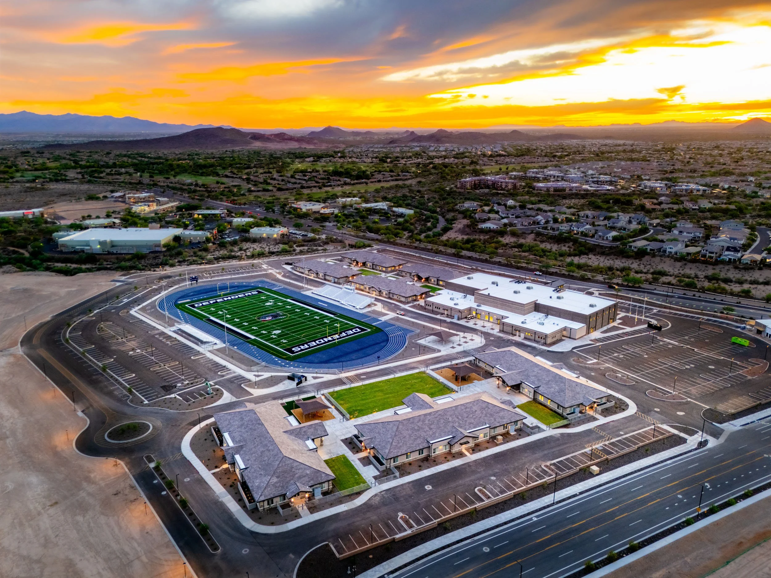 Aerial view of a sports complex at sunset with a football field, parking lots, and surrounding buildings in a desert landscape.