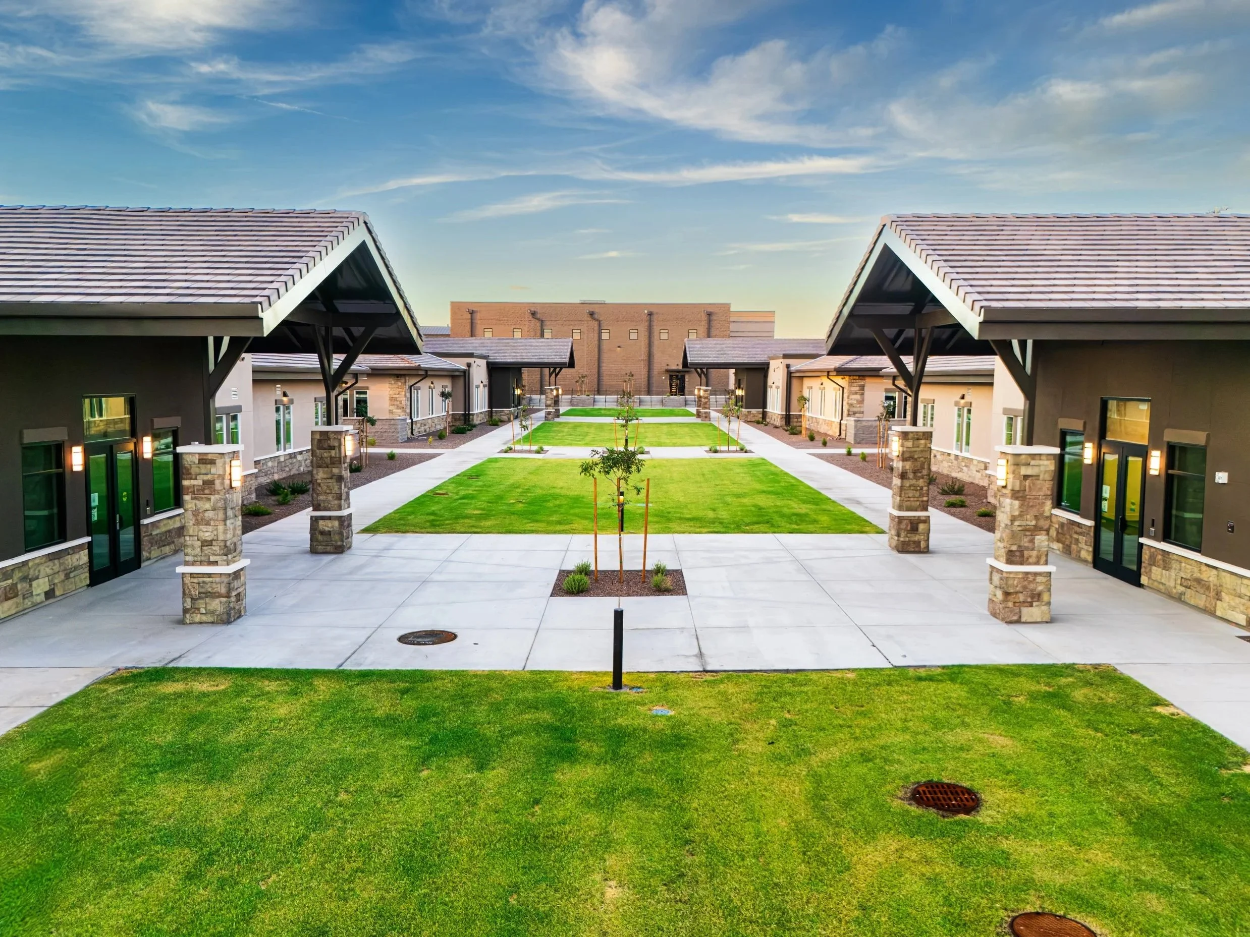A modern apartment complex courtyard with green grass, paved walkways, and townhouses with stone accents and large windows under a partly cloudy sky.