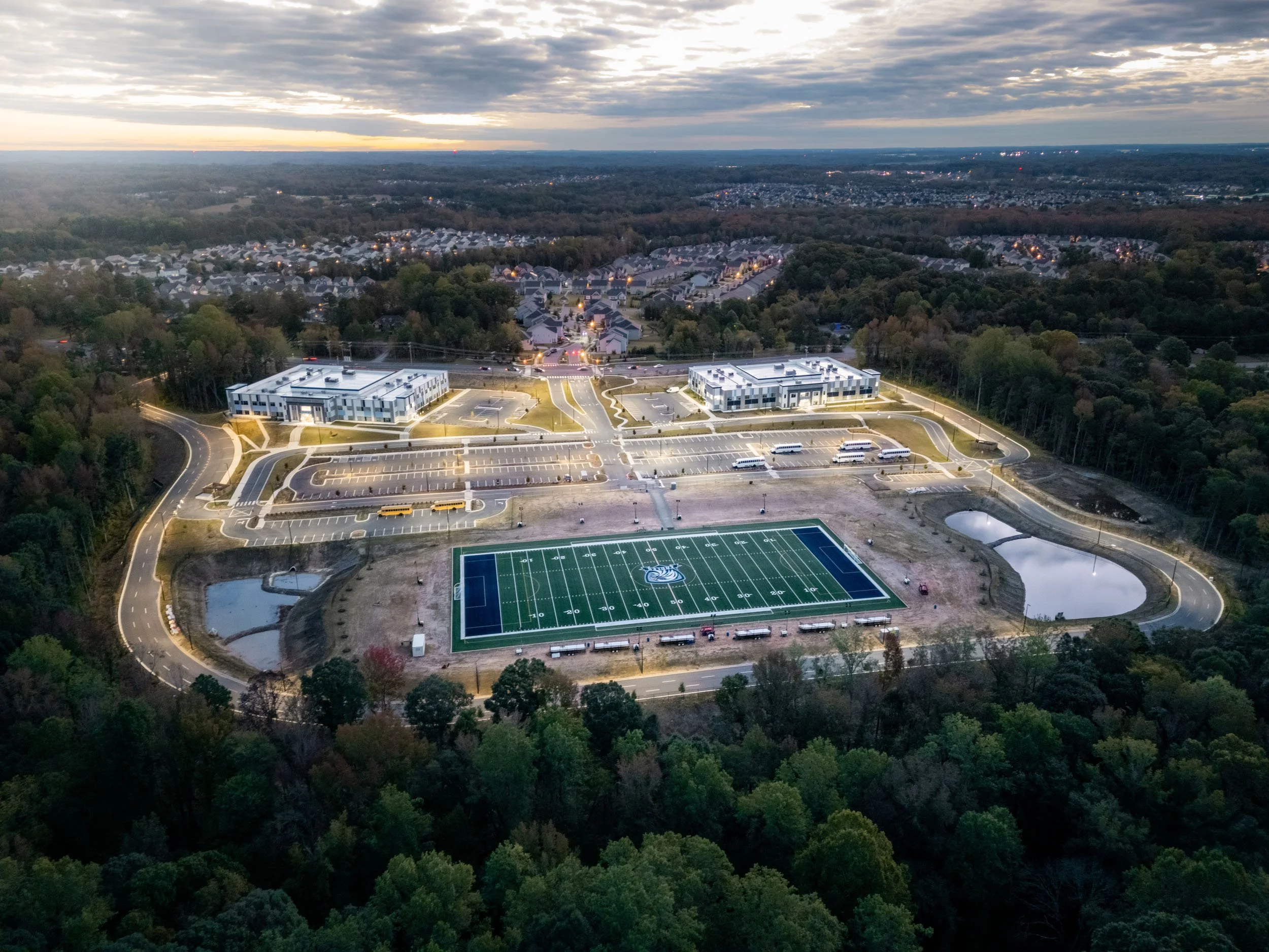 Aerial view of a school campus with a football field, parking lots, and two modern buildings, surrounded by trees, at dusk.