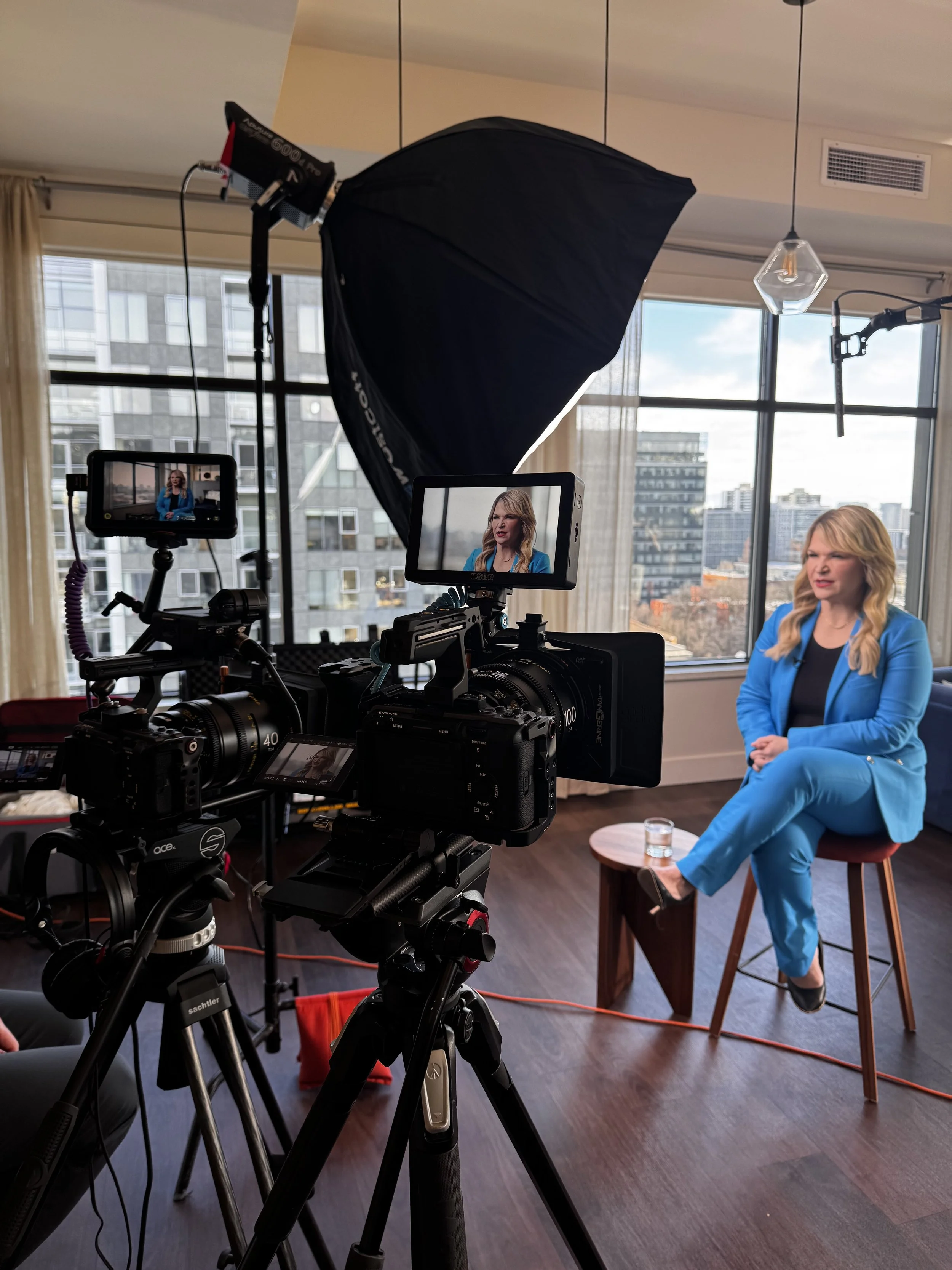 A woman in a blue suit sitting on a chair during an interview in a room with large windows and city view, with professional lighting and camera equipment filming her.