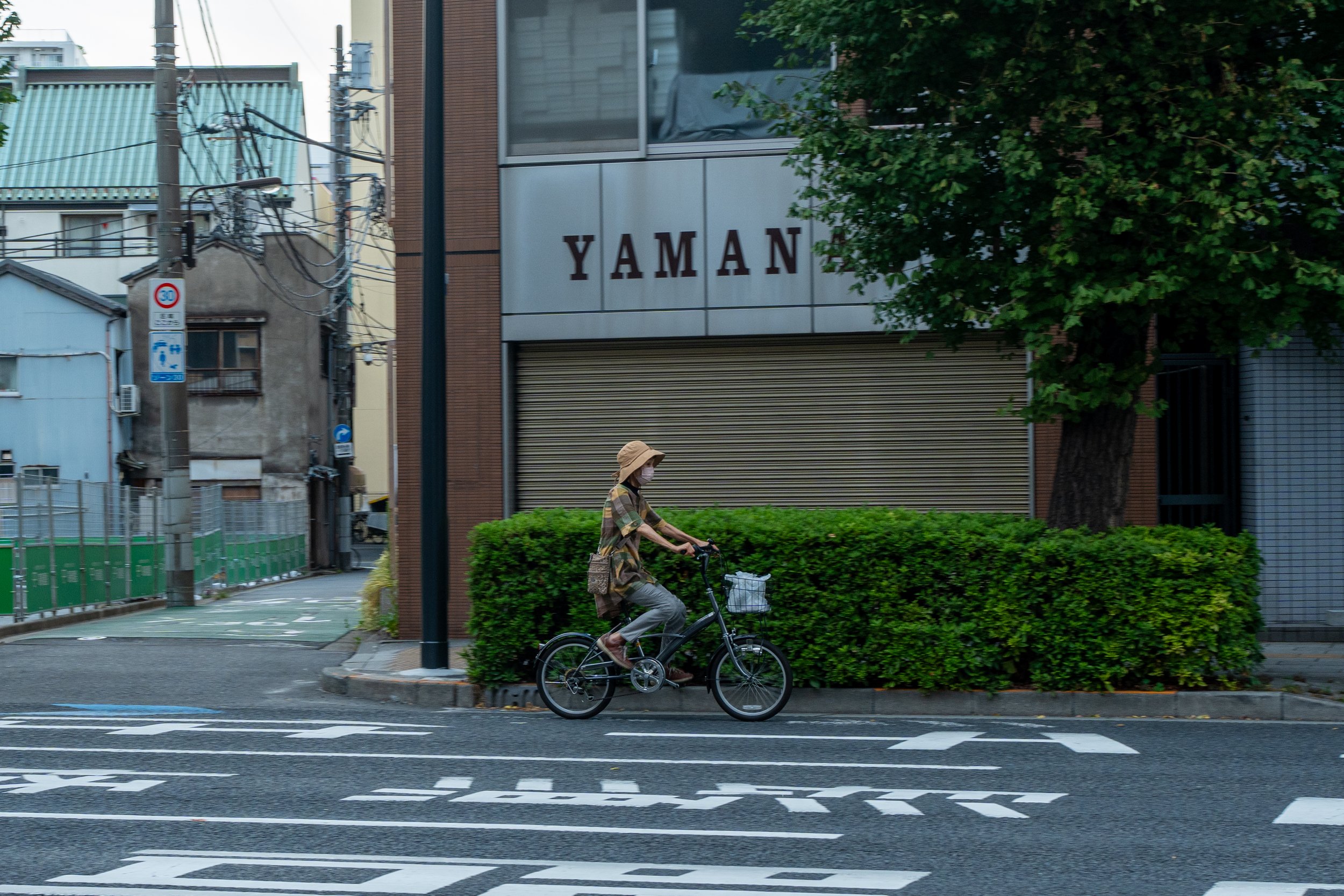 A woman cyclist riding a bicycle on a city street, wearing a hat, face mask, and casual clothes, with buildings and greenery in the background.
