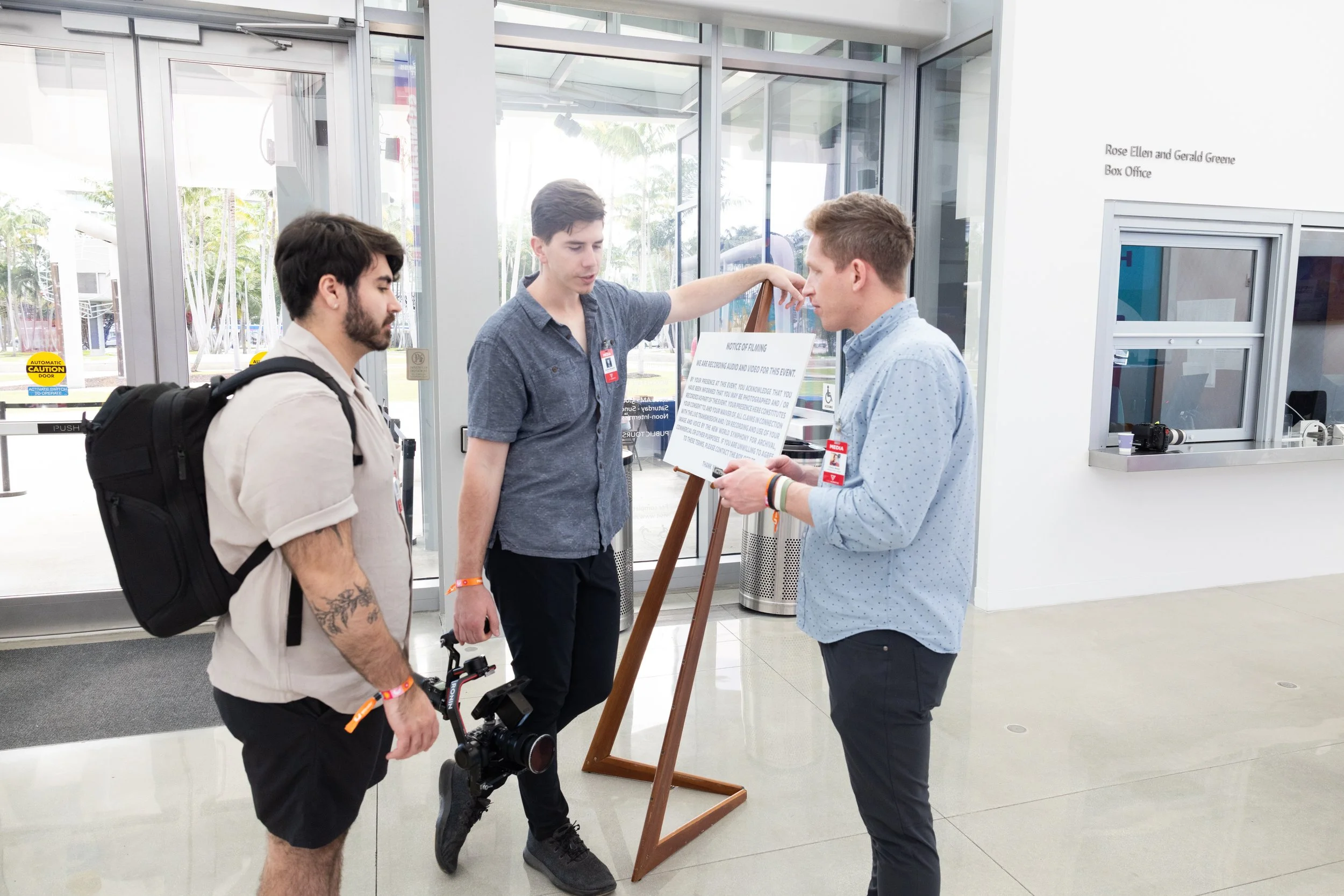 Three men standing near an entrance, two of them looking at a notice board held by a third man. The man on the left has a backpack and tattoos on his arm, the man in the center is wearing a short-sleeved shirt and has a camera hanging from his neck, 