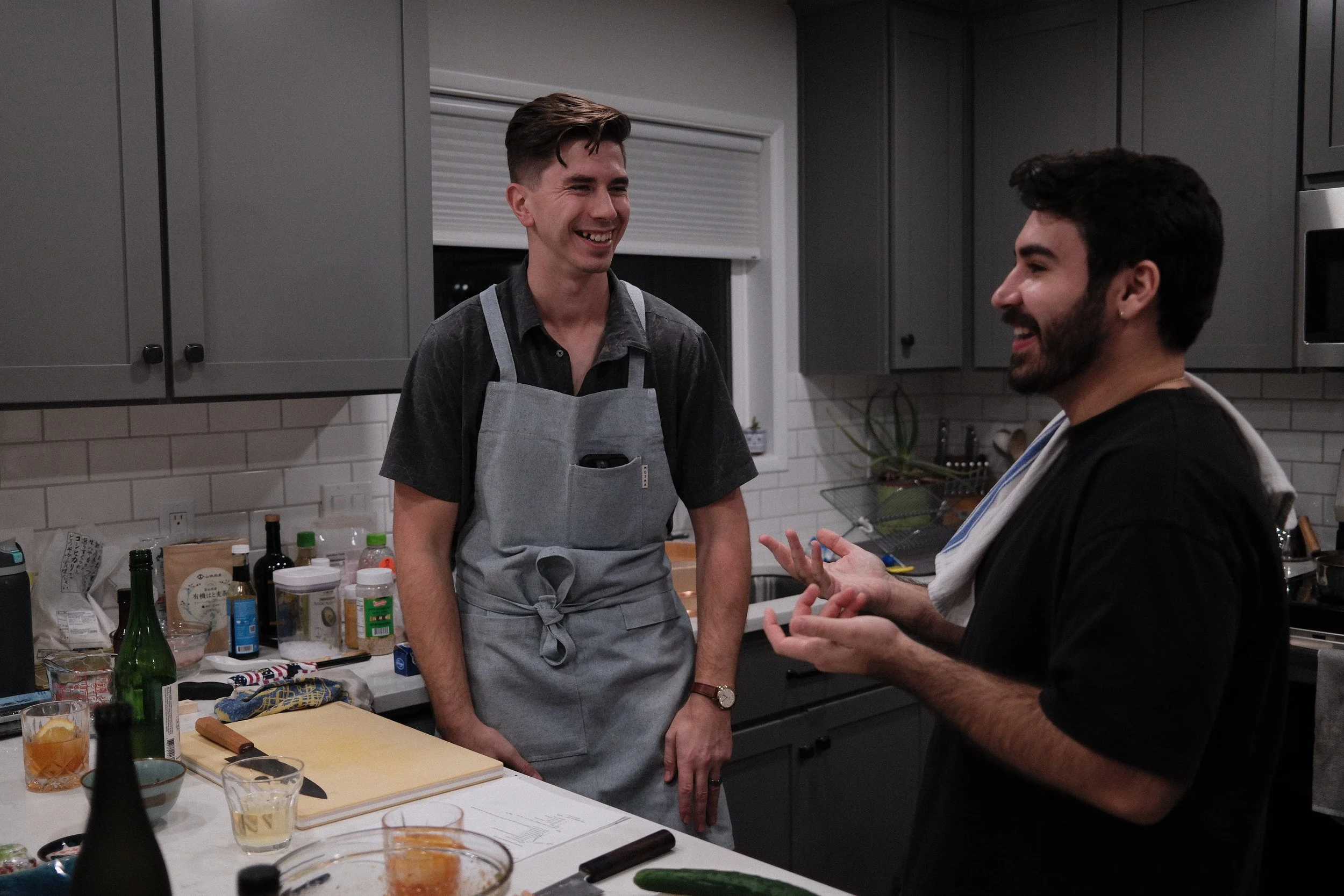 Two men are in a kitchen smiling and talking. One man is wearing a gray apron, and the other is dressed casually. The kitchen counter is cluttered with bottles, jars, a cutting board, and glasses.