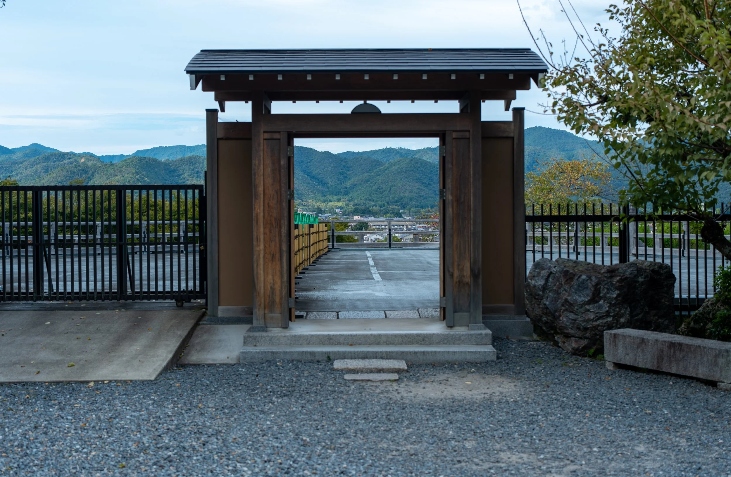 Traditional Japanese wooden gate overlooking a parking lot and distant green mountains under a partly cloudy sky.