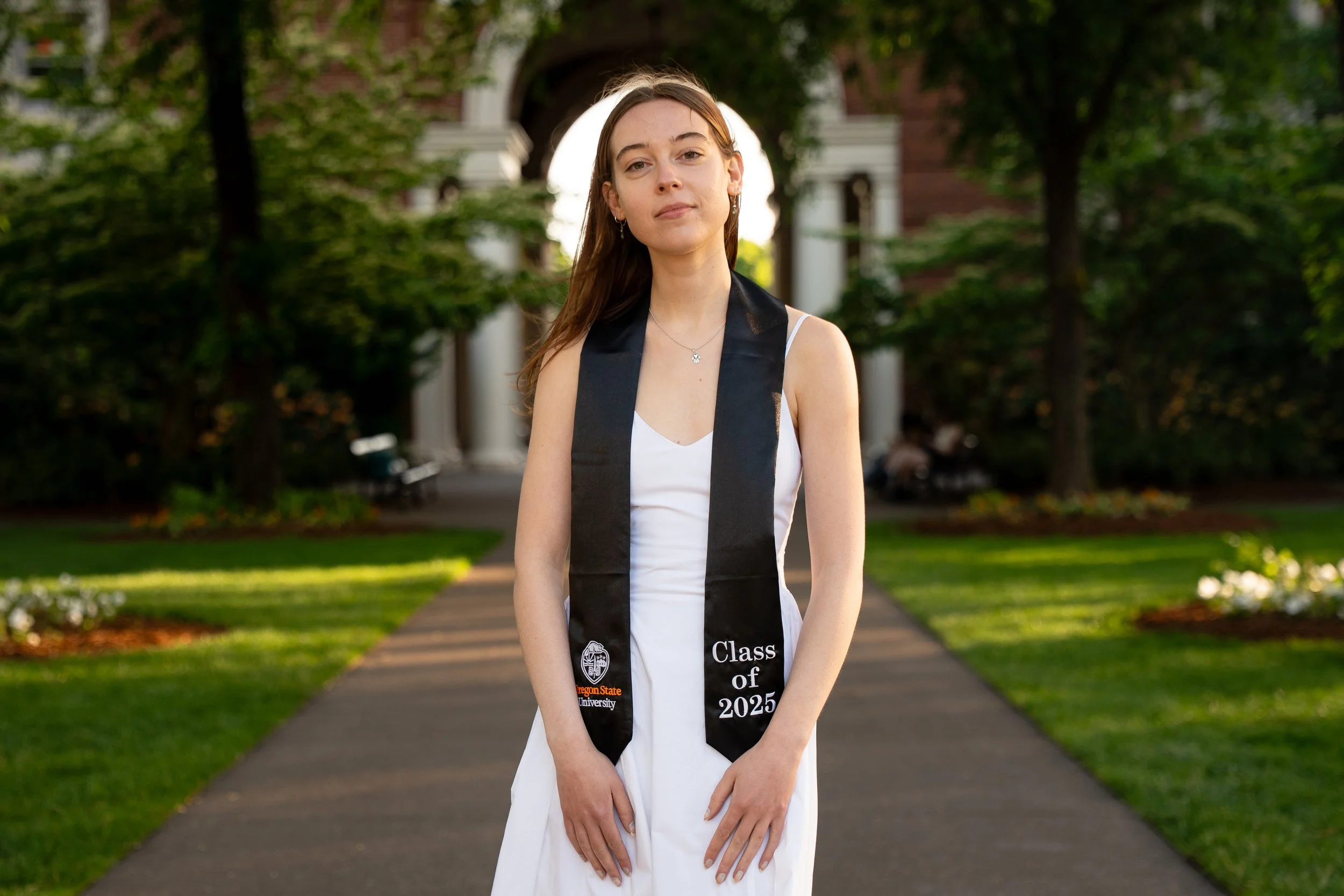 A young woman in a white dress wearing a black graduation stole that reads 'Class of 2025' and features the Oregon State University logo, standing on a pathway outdoors with greenery and trees in the background.