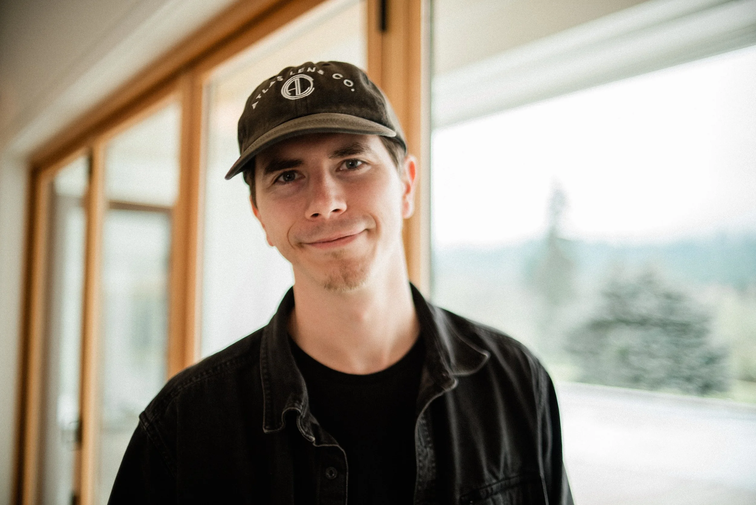 A young man with short dark hair, wearing a black baseball cap and black jacket, standing indoors near a large window with a scenic view outside.