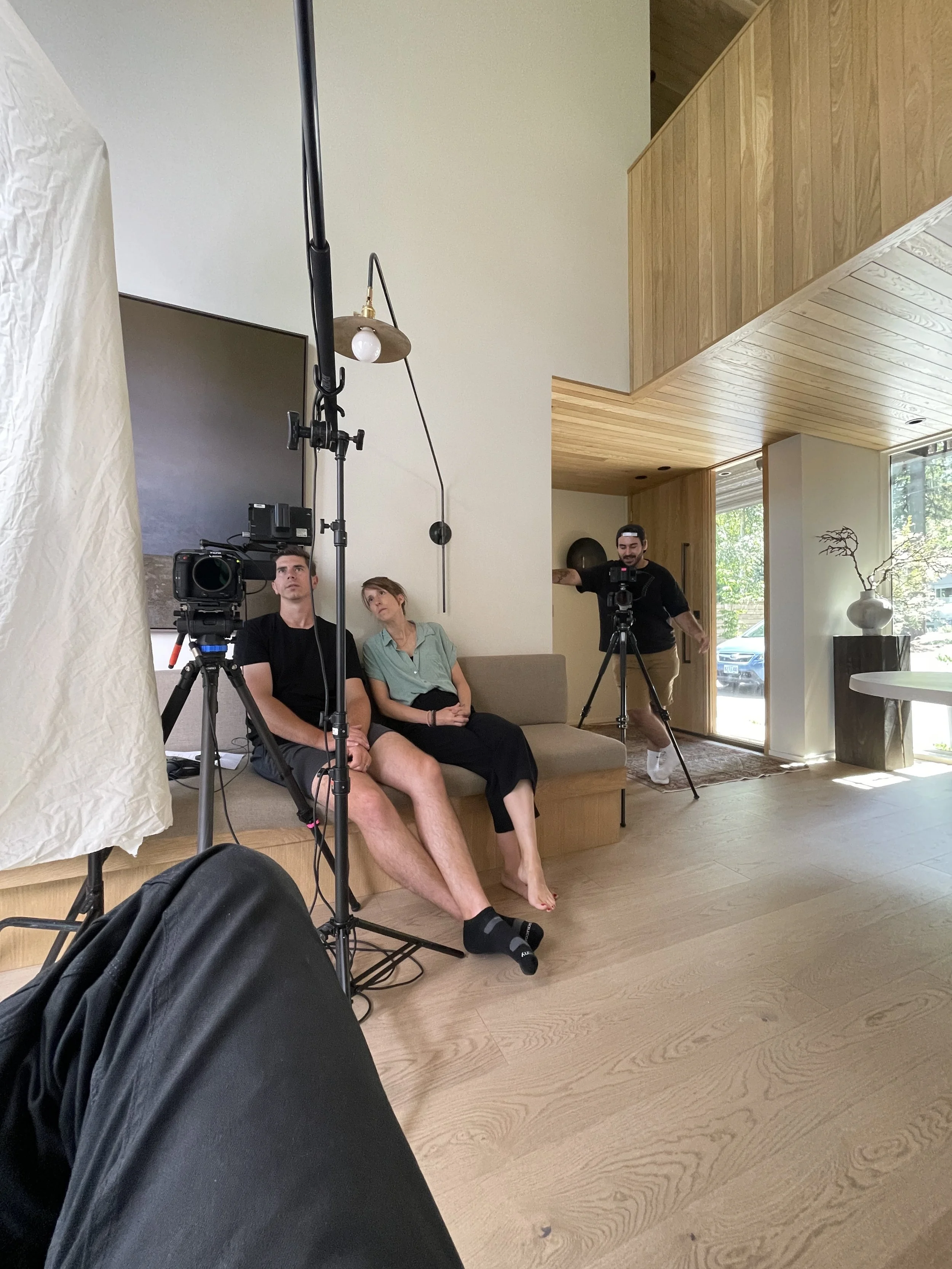 Two people sitting on a sofa watching a video shoot, with a person adjusting camera equipment behind them, inside a modern house with wooden accents and large windows.