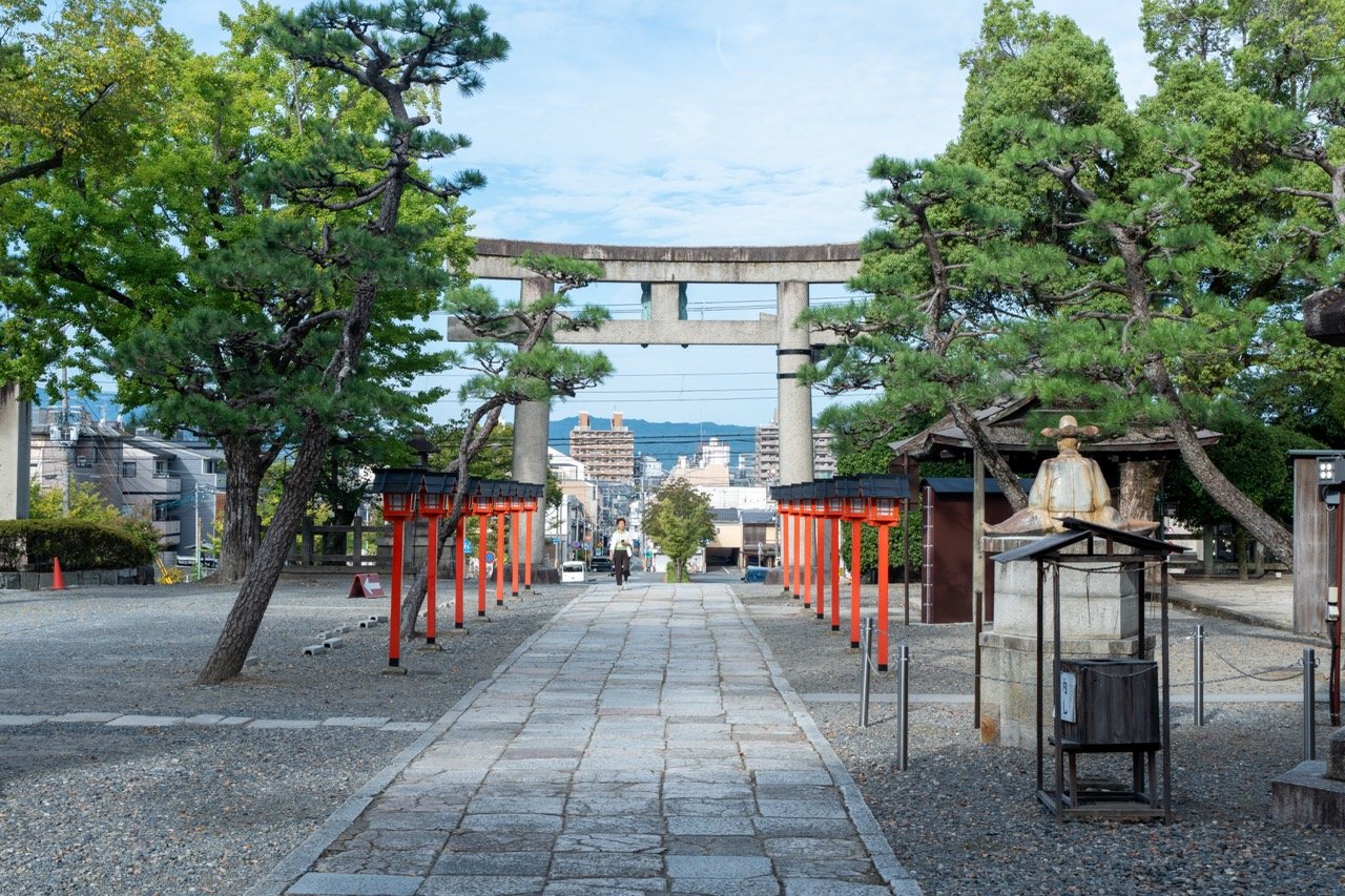 A Japanese shrine entrance with a stone pathway leading to a torii gate, surrounded by green trees, red lanterns, and traditional statues.