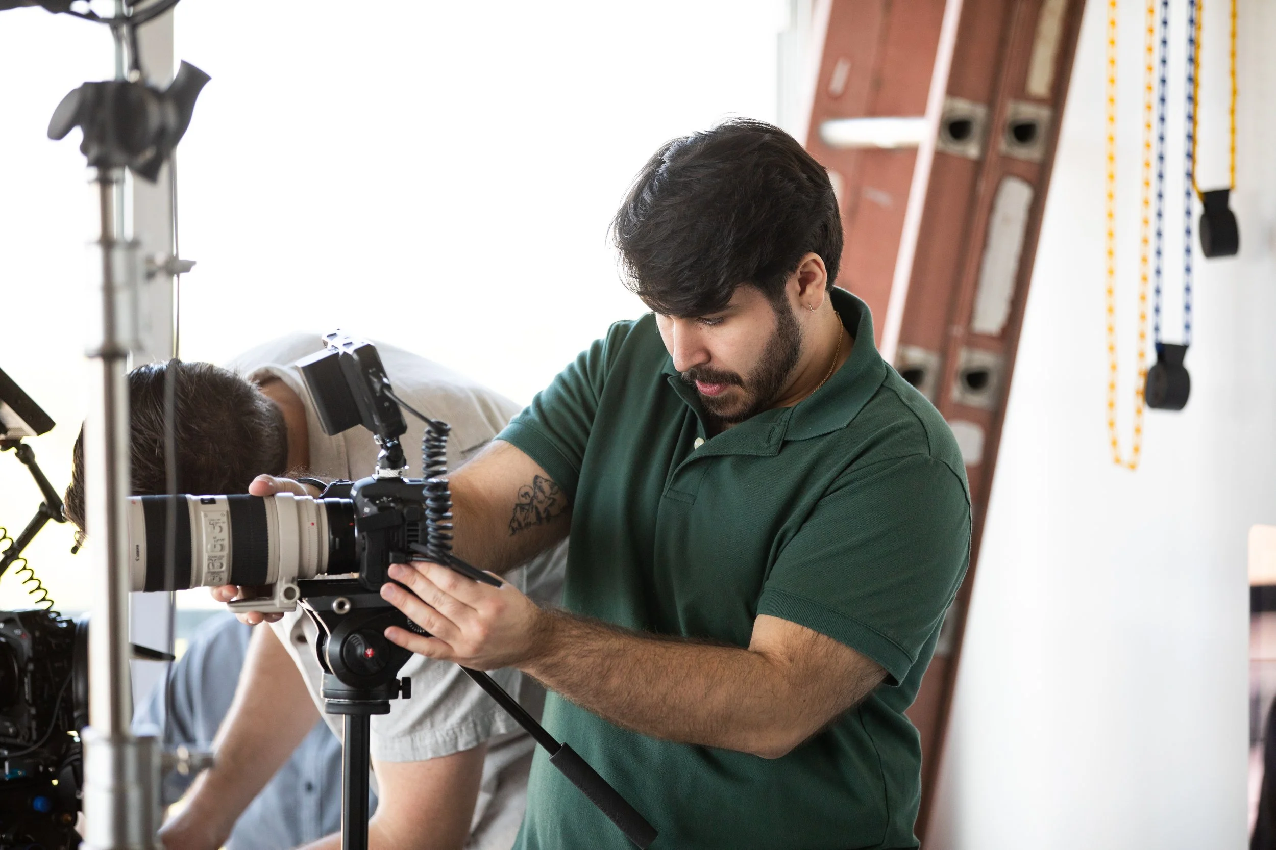 A man with dark hair and a beard adjusting a professional camera on a tripod during a photoshoot or filming session.