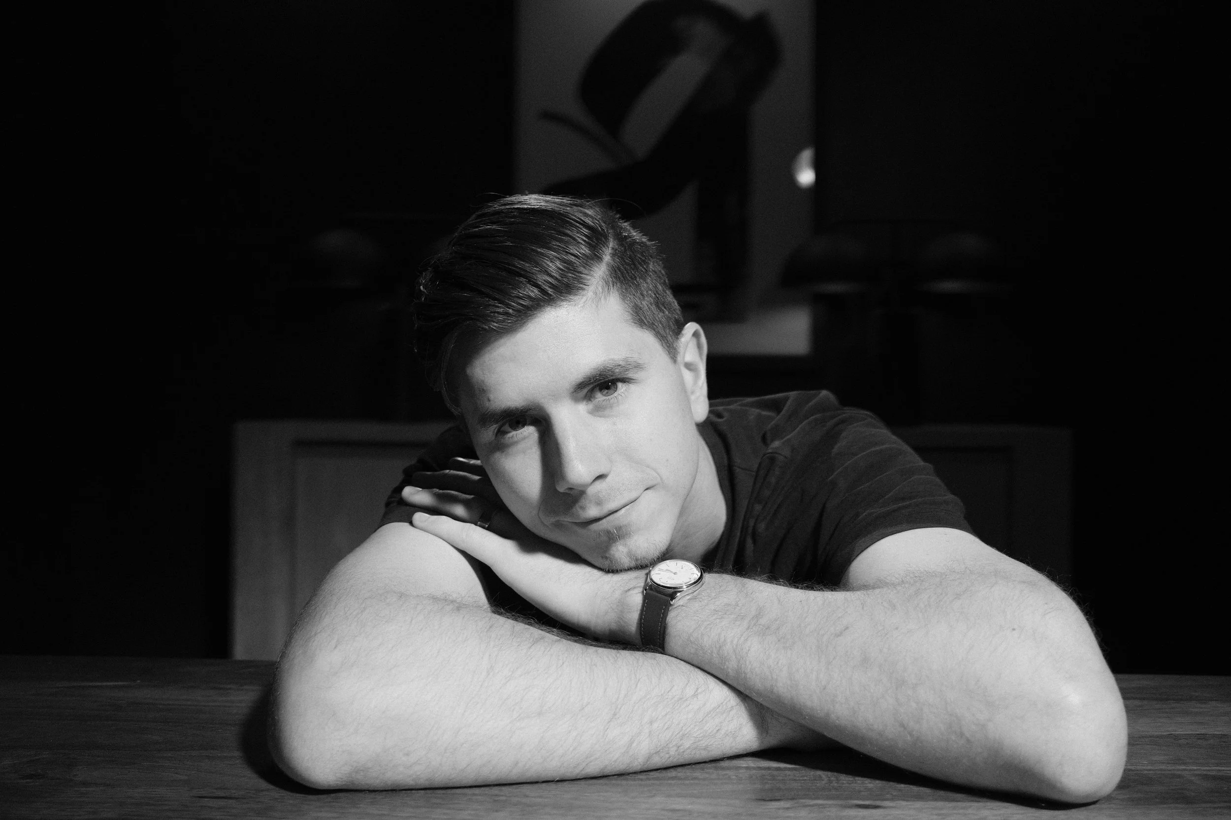 Black and white photo of a young man with short hair, leaning on a table, resting his head on his crossed arms, smiling softly at the camera.