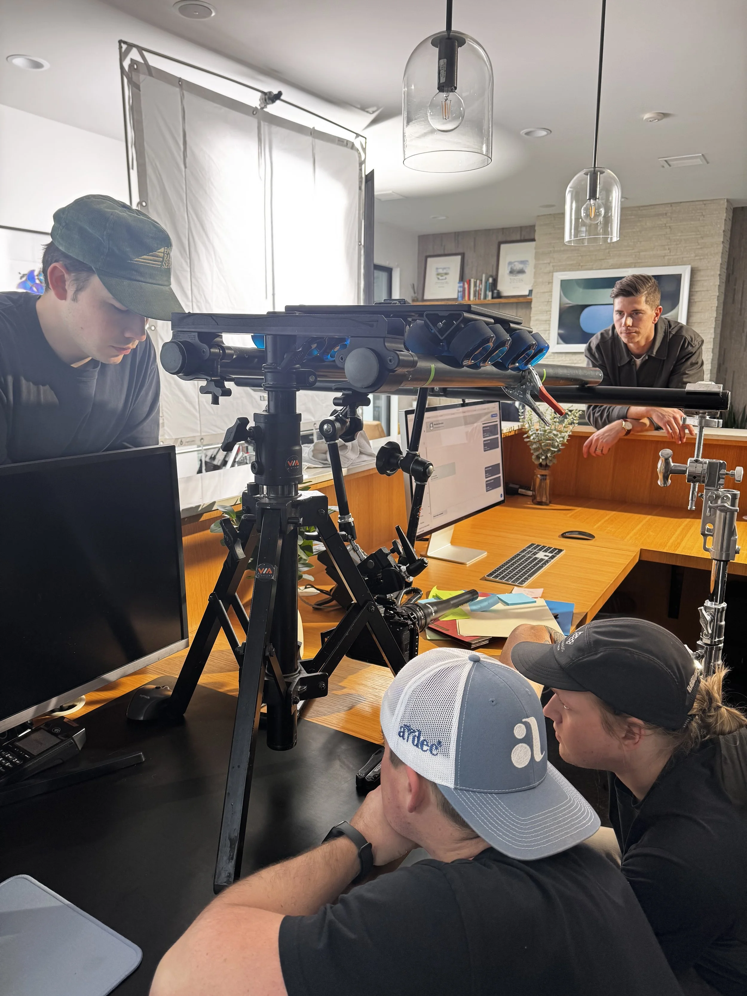A group of people working on a film or photography shoot in an indoor office setting, with cameras, monitors, notebooks, and lighting equipment.