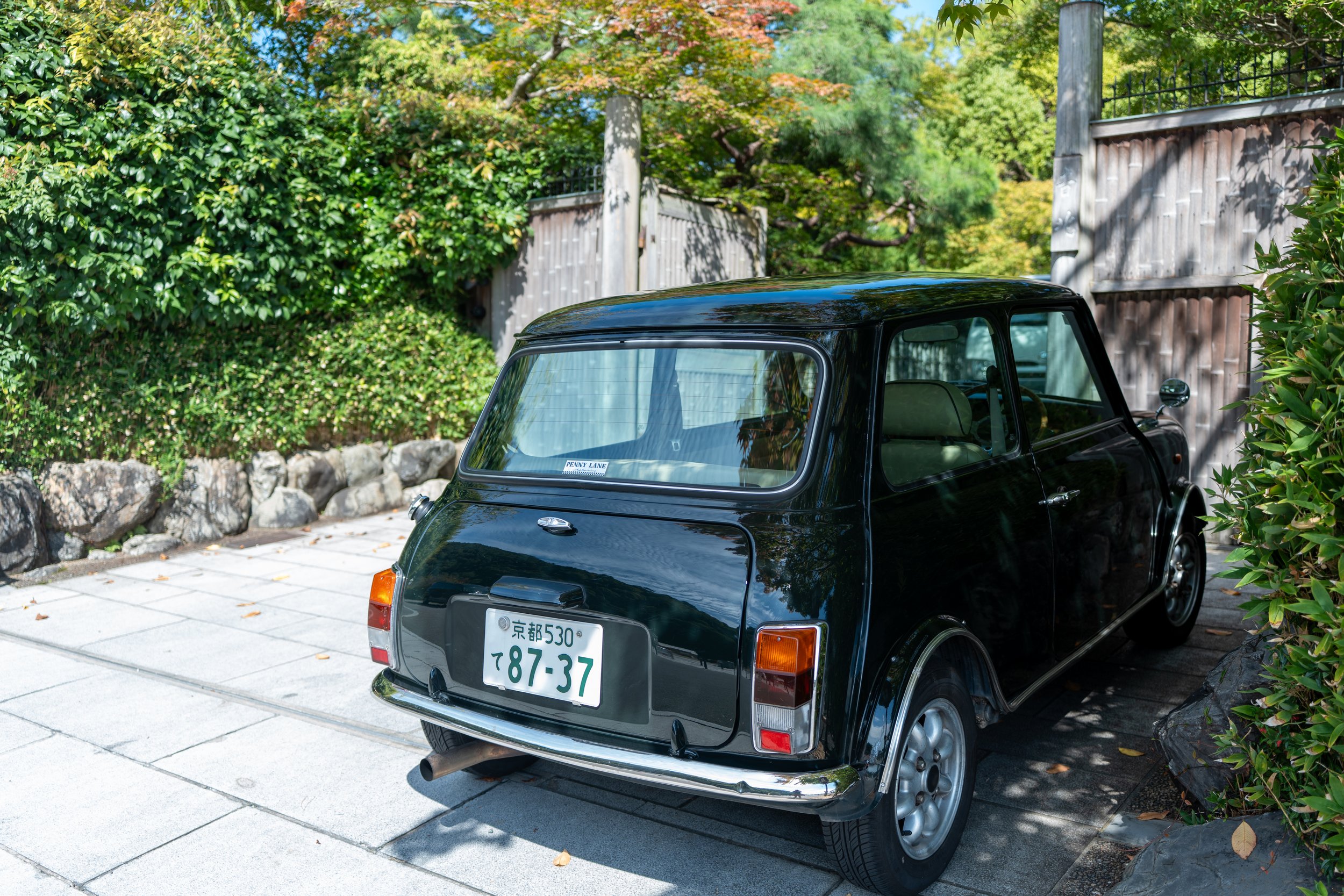 A vintage black microcar parked on a paved area next to green bushes and stone landscaping, with trees and a wooden fence in the background.