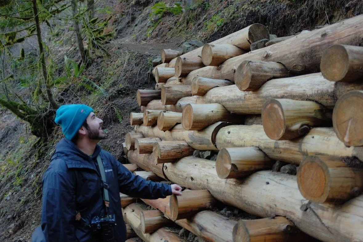 A man in outdoor clothing and a blue beanie stands next to a large stack of cut logs in a forested area.