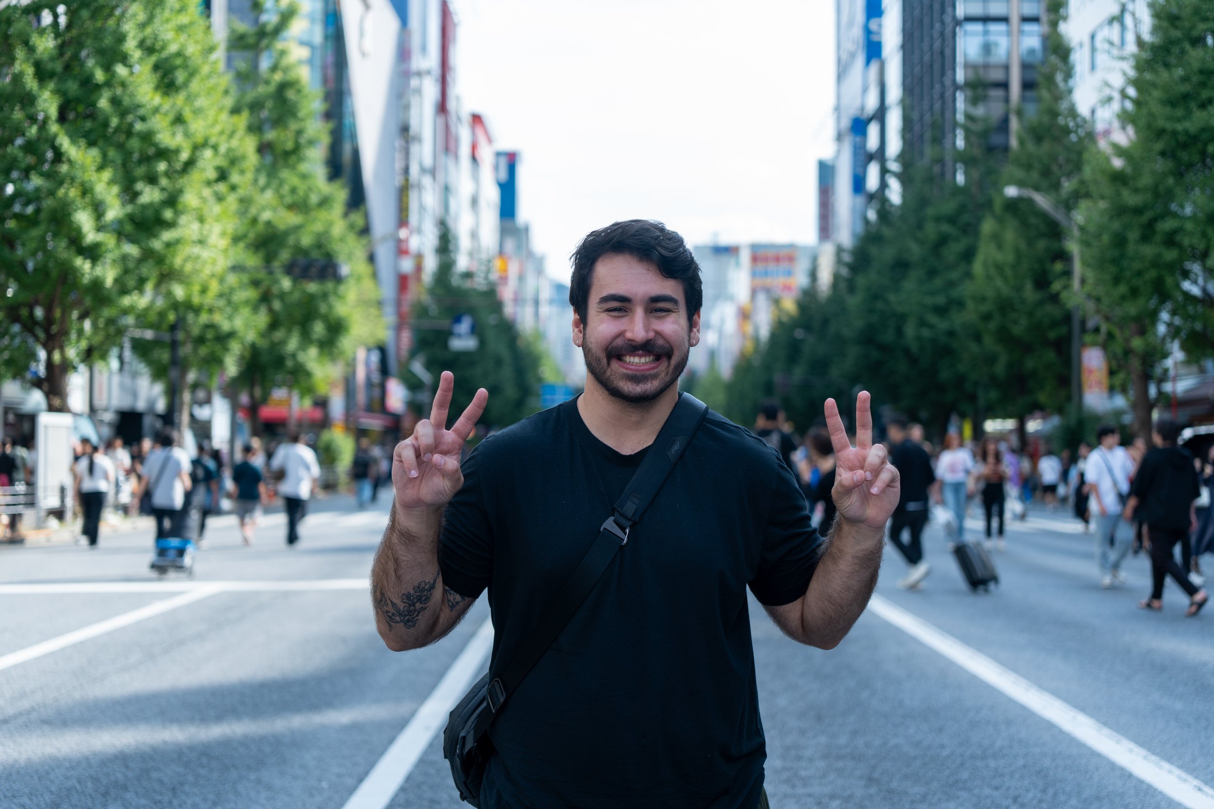 Smiling man with a beard and black shirt making peace signs with both hands on a busy city street with pedestrians and tall buildings.